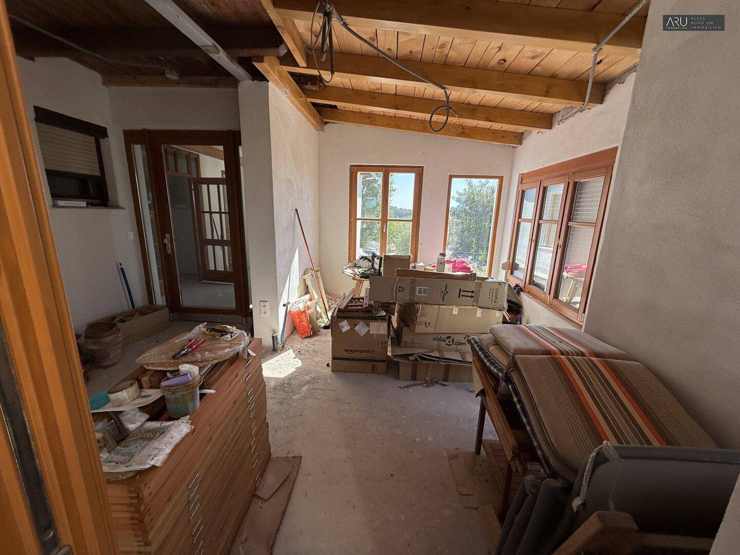 Innenraum im Rohbau mit Holzbalkendecke und Blick durch die Fenster ins Grüne.