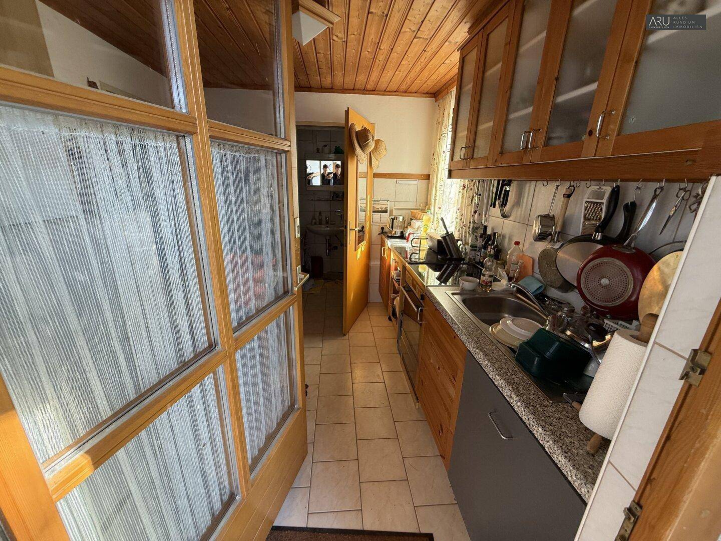 Schmale Küche mit Holzschränken, Fliesenboden und Blick nach draußen.