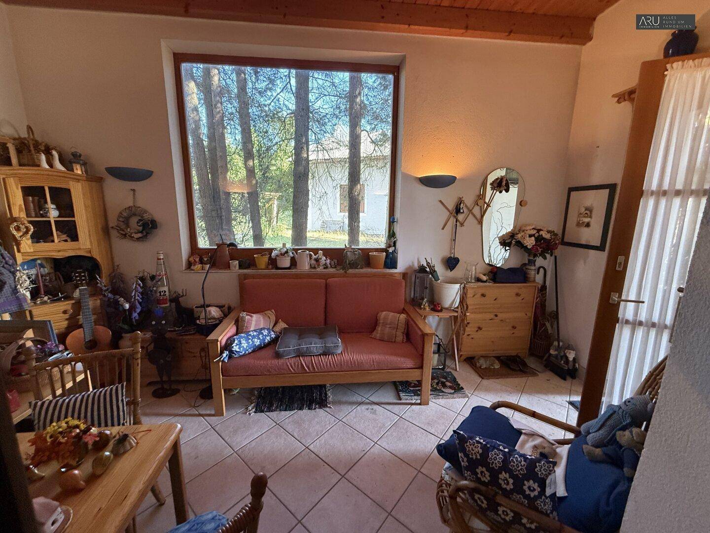 Wohnzimmer mit Fliesenboden, großem Fenster und rustikaler Holzmöblierung.