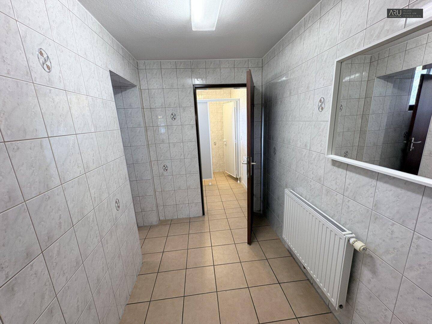 Gefliester Flur mit Heizkörper und Spiegel, führt zu weiteren Sanitärräumen.