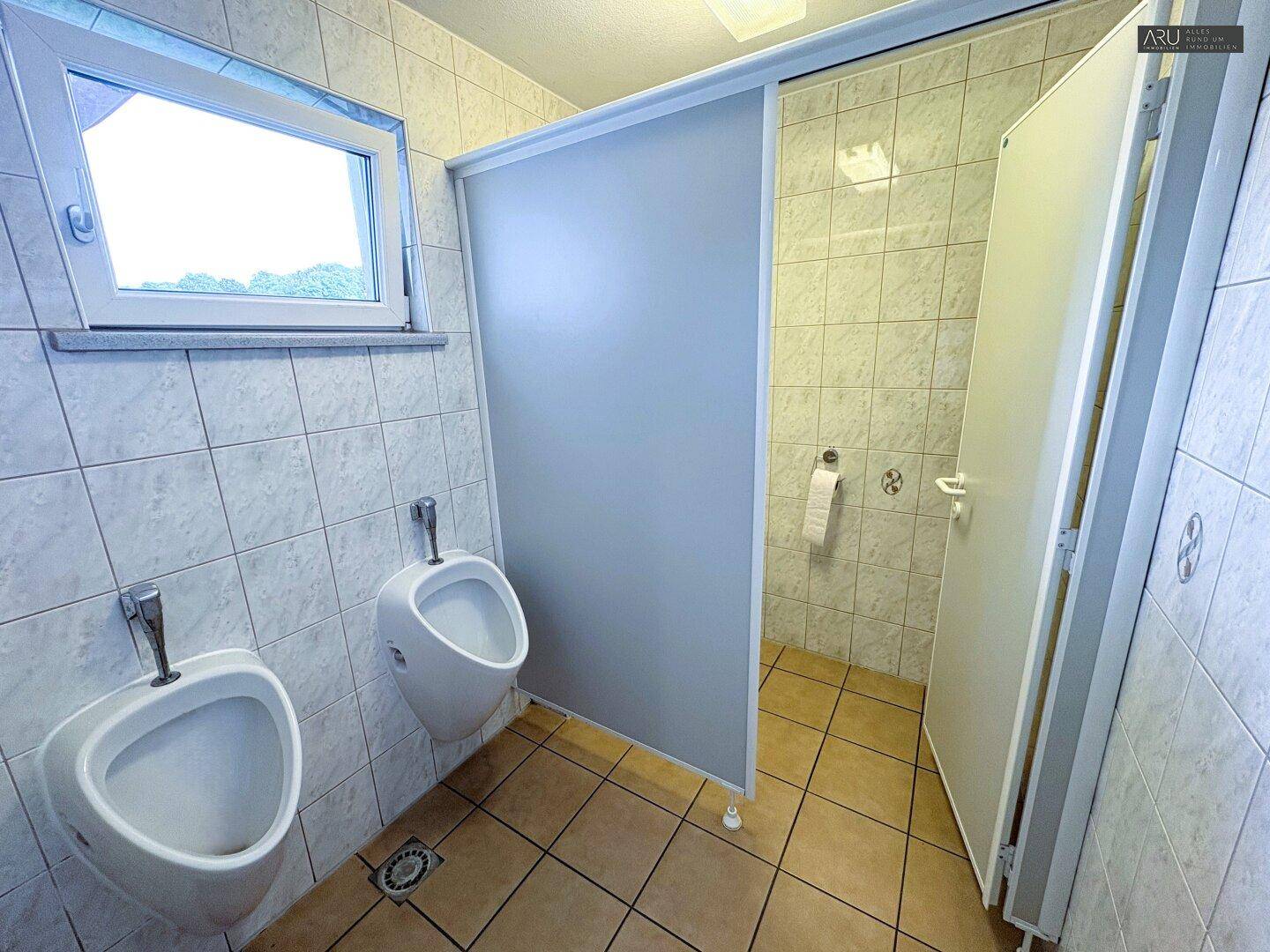Herren-WC mit zwei Urinalen und einer Toilettenkabine, ausgestattet mit Fenster und Fliesen.