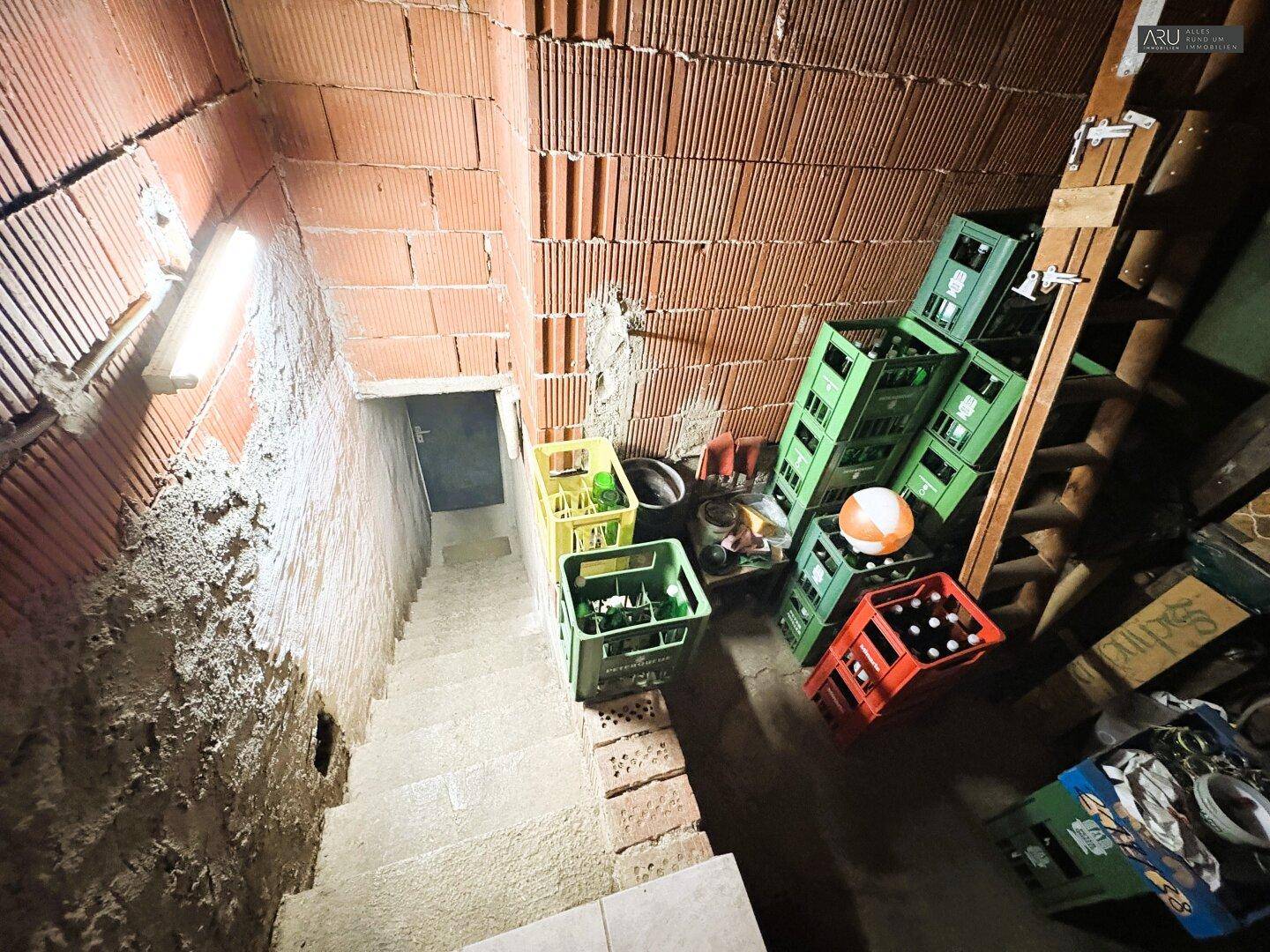Treppe führt hinab in einen Kellerraum mit unverputzten Ziegelwänden und Lagerkisten.