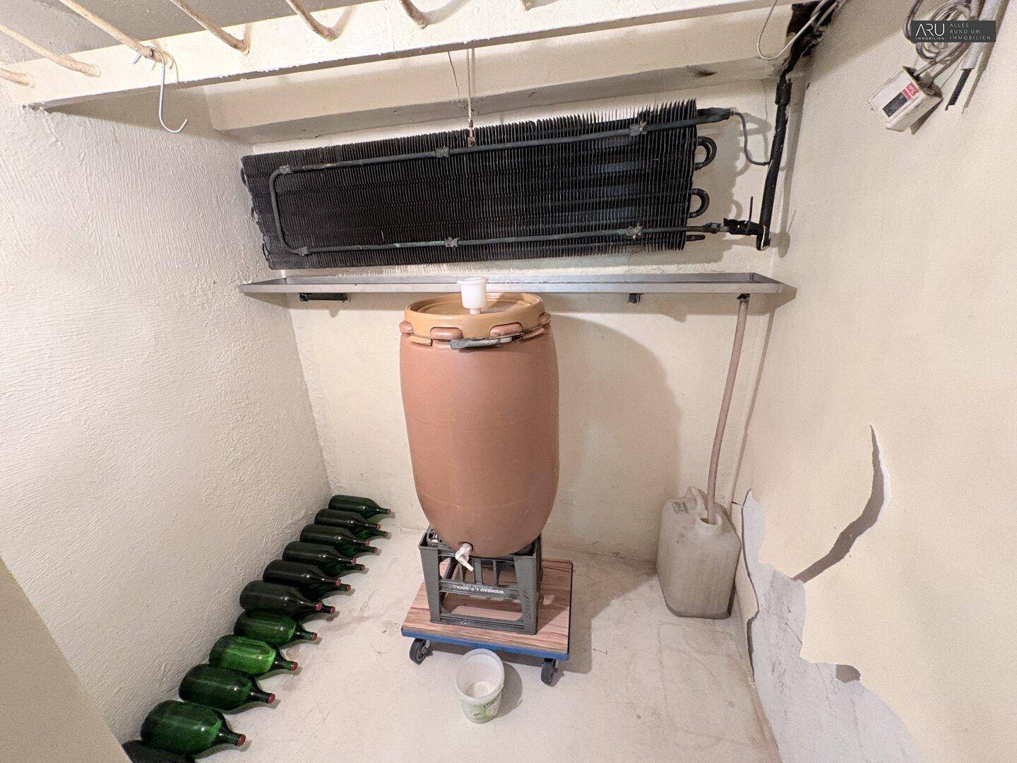 Kleiner Weinkeller mit gelagerten Flaschen und einem großen Gärbehälter.