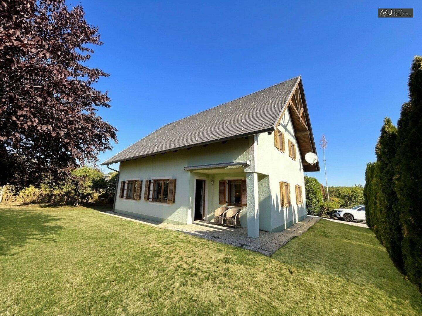 Das Einfamilienhaus ist von einem weitläufigen Garten mit Rasenflächen und Bäumen umgeben, unter blauem Himmel.