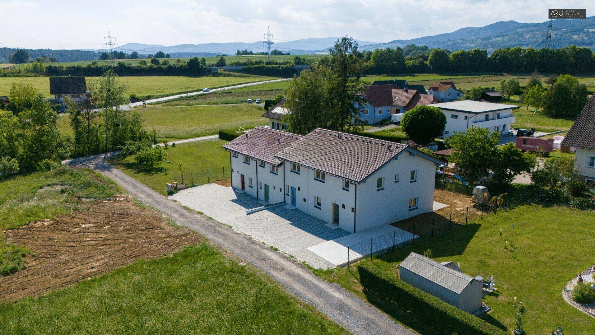 Weitläufige Luftaufnahme der Reihenhäuser in ländlicher Umgebung mit Feldern und Bergen im Hintergrund.