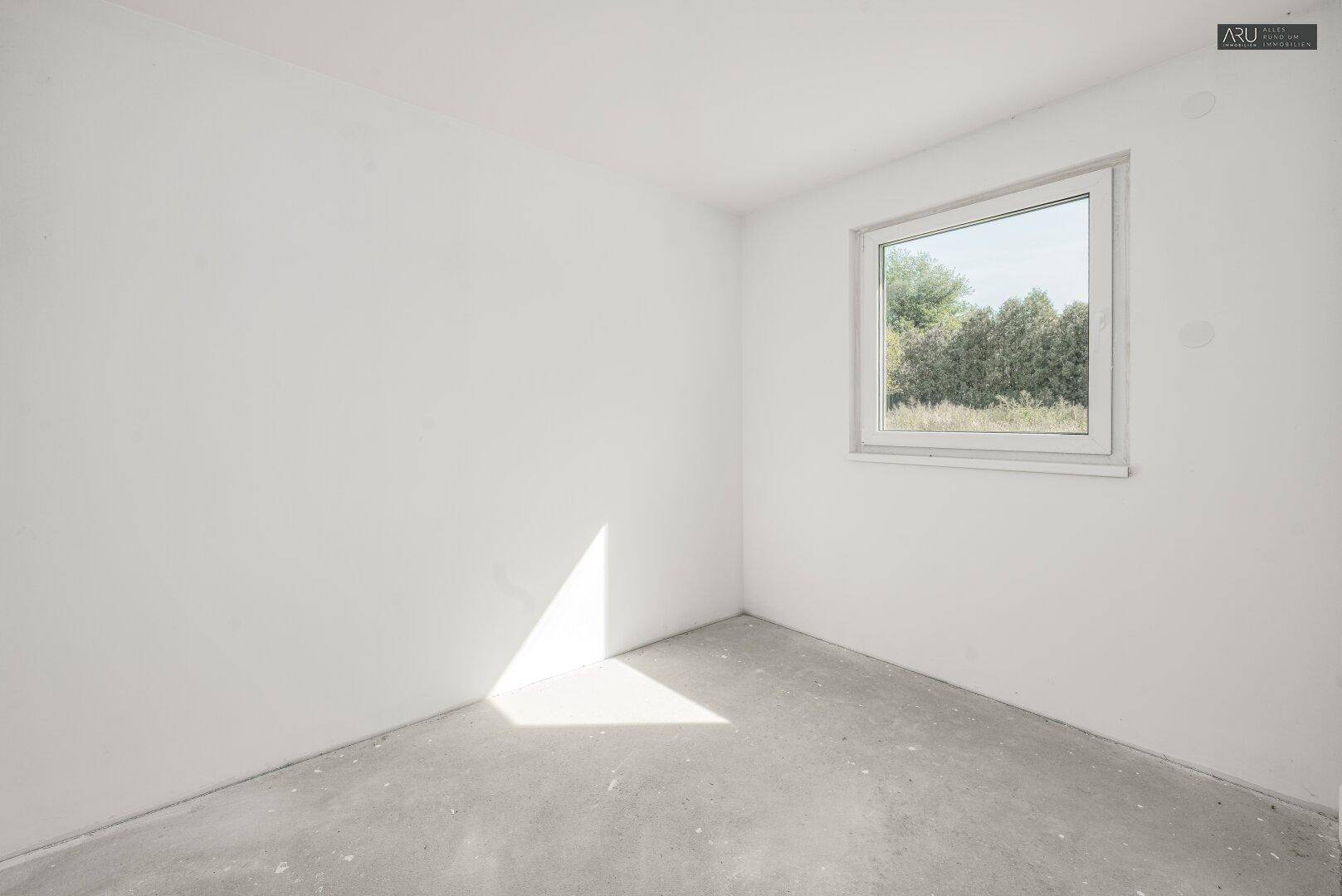 Helles, unmöbliertes Zimmer mit weiß gestrichenen Wänden, Betonboden und Fenster mit Grünblick.