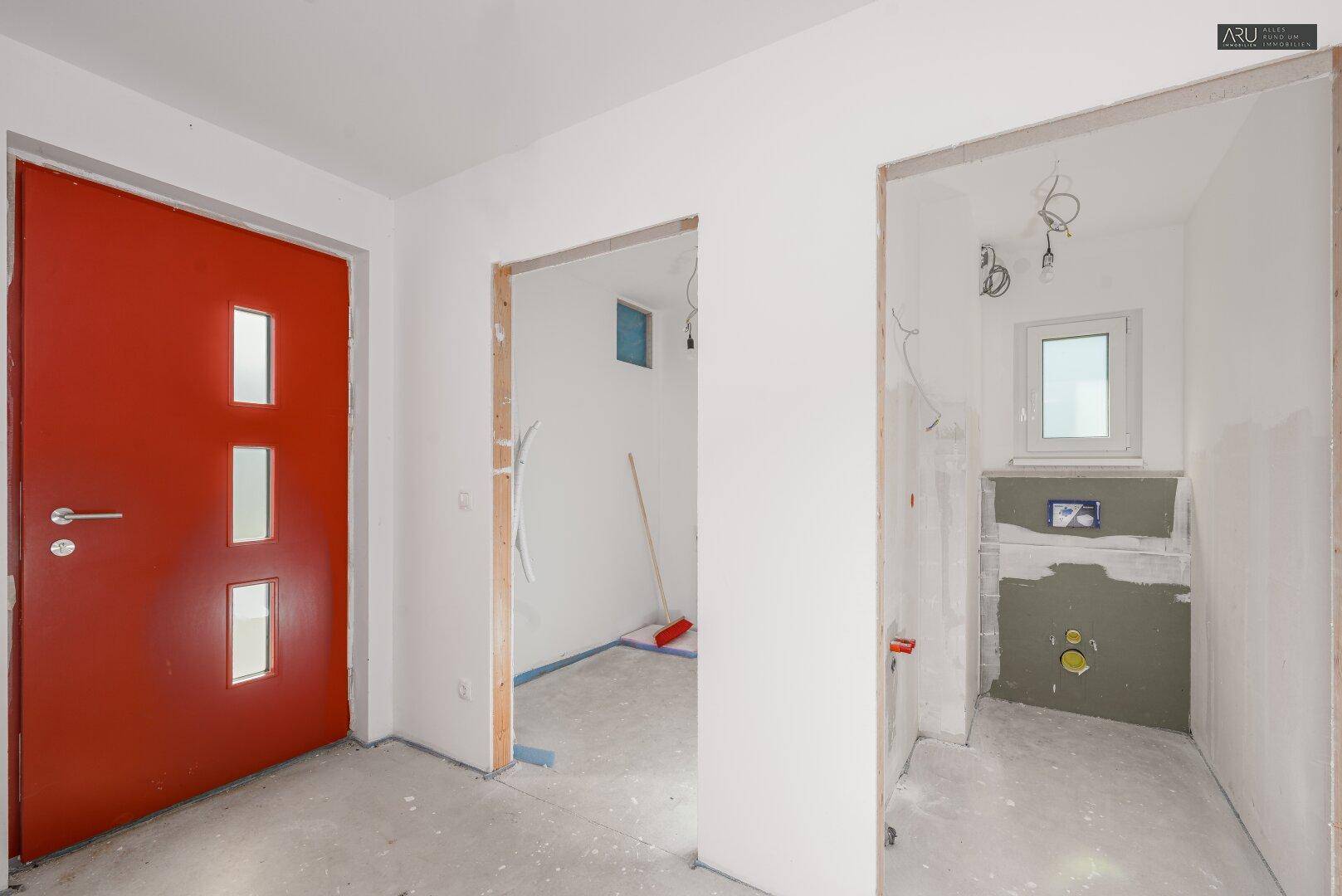 Eingangsbereich mit roter Haustür, Betonboden und Zugängen zu weiteren unfertigen Räumen.