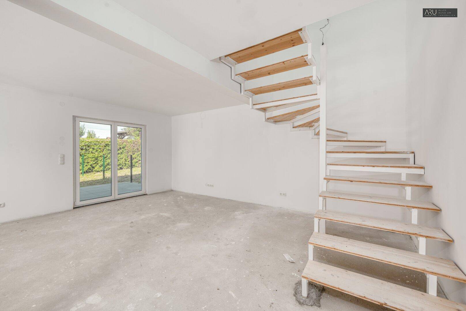 Geräumiger, unmöblierter Raum mit unfertiger Holztreppe, Schiebetür und Blick ins Grüne.