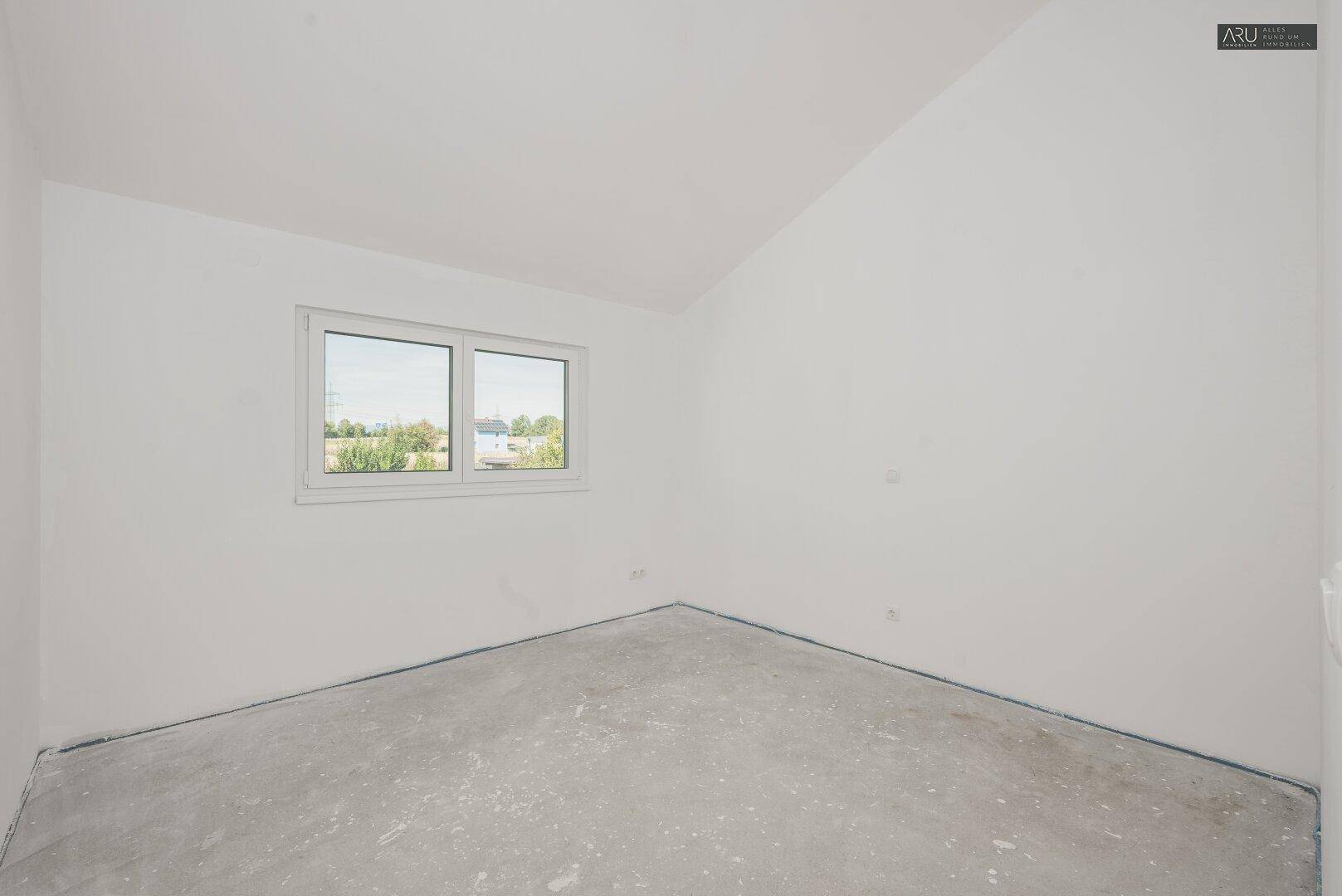 Helles, unmöbliertes Zimmer im Dachgeschoss mit Betonboden, Fenster und weitem Ausblick.
