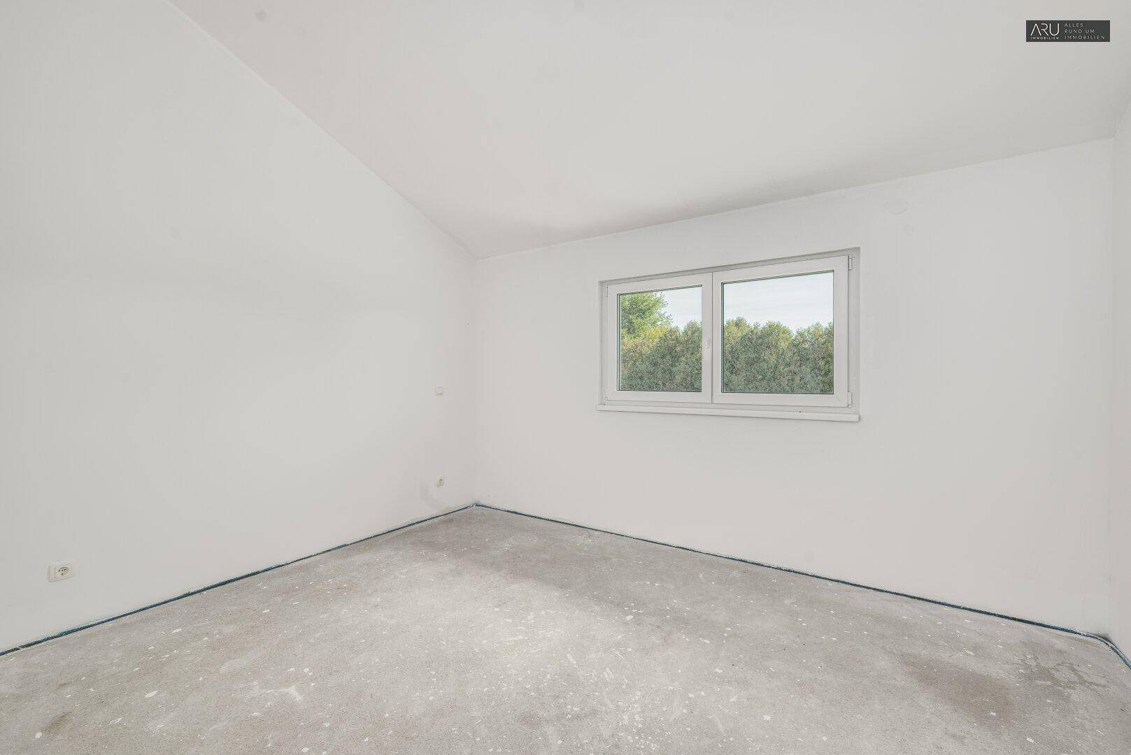 Geräumiges, unmöbliertes Zimmer im Dachgeschoss mit Betonboden, großem Fenster und schräger Decke.