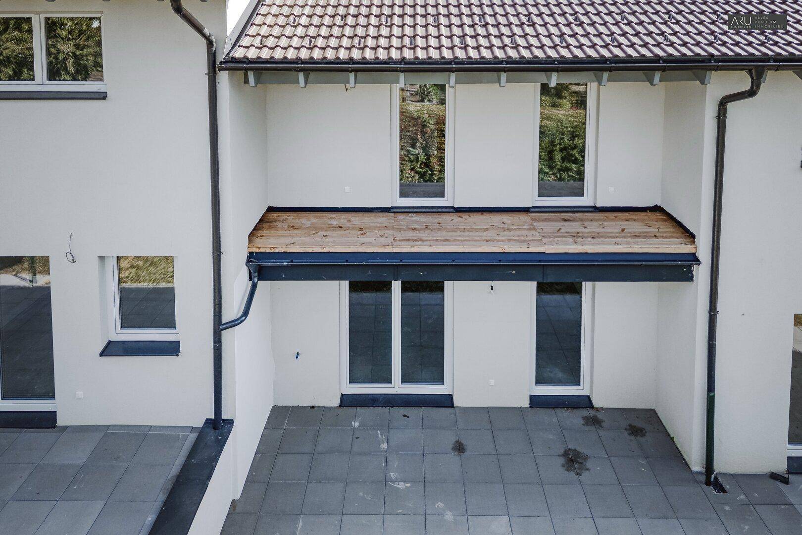 Außenansicht mit großer gepflasterter Terrasse und darüberliegendem Holzbalkon.