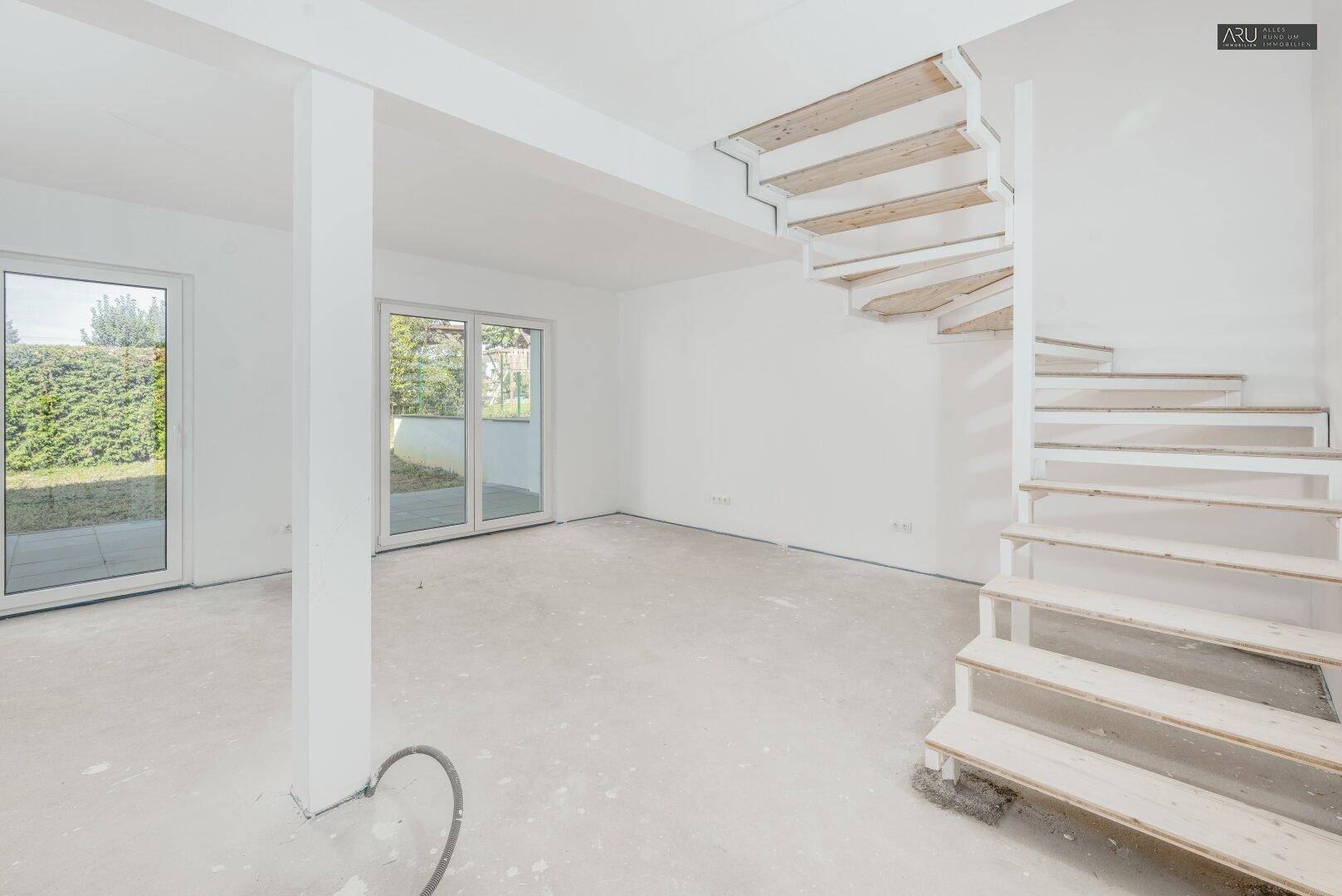 Heller, unfertiger Wohnbereich mit Rohbau-Treppe und Zugang zum Garten durch große Terrassentüren.
