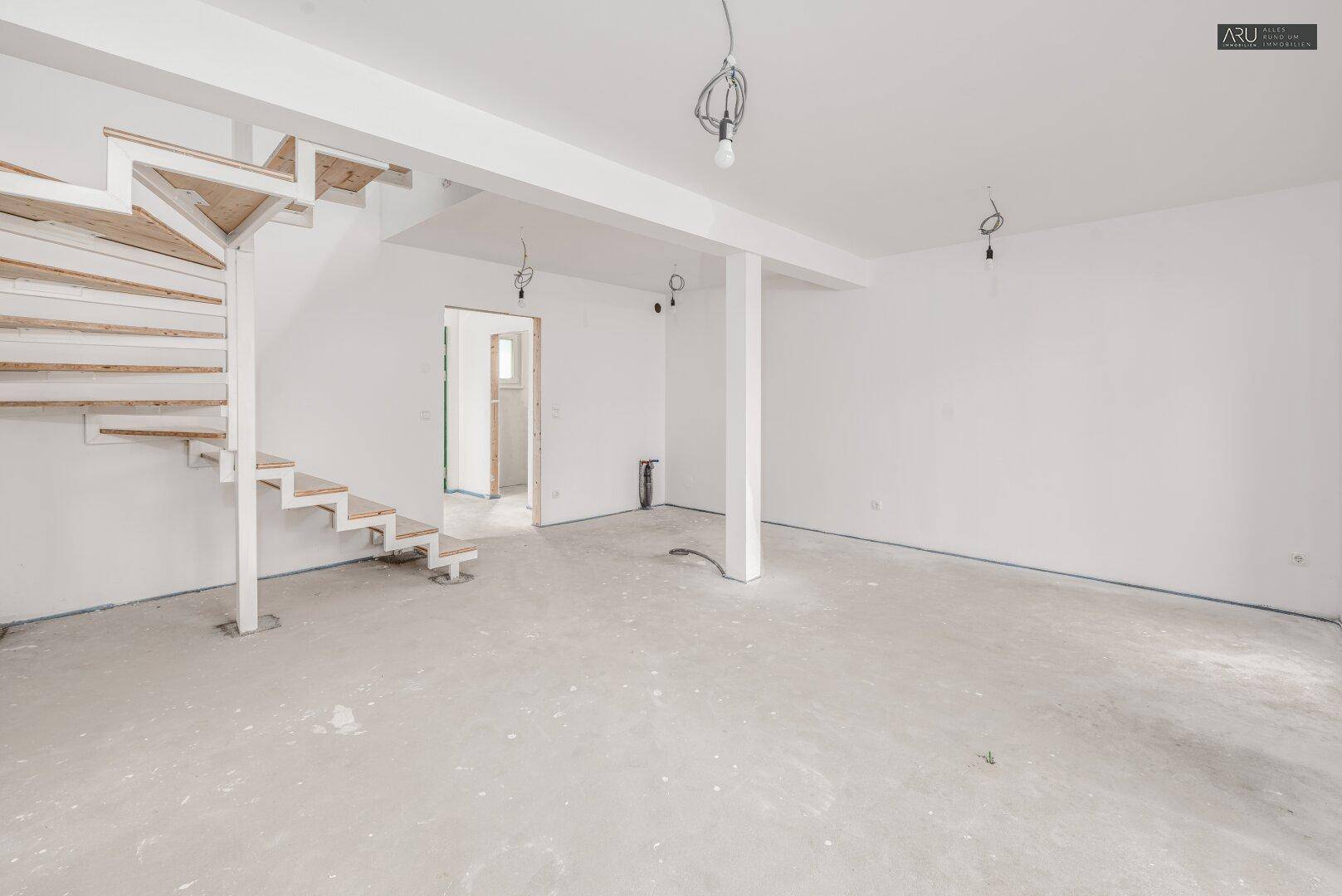 Geräumiger, unfertiger Wohnbereich mit offener Treppe und Säulen, bereit für den Innenausbau.