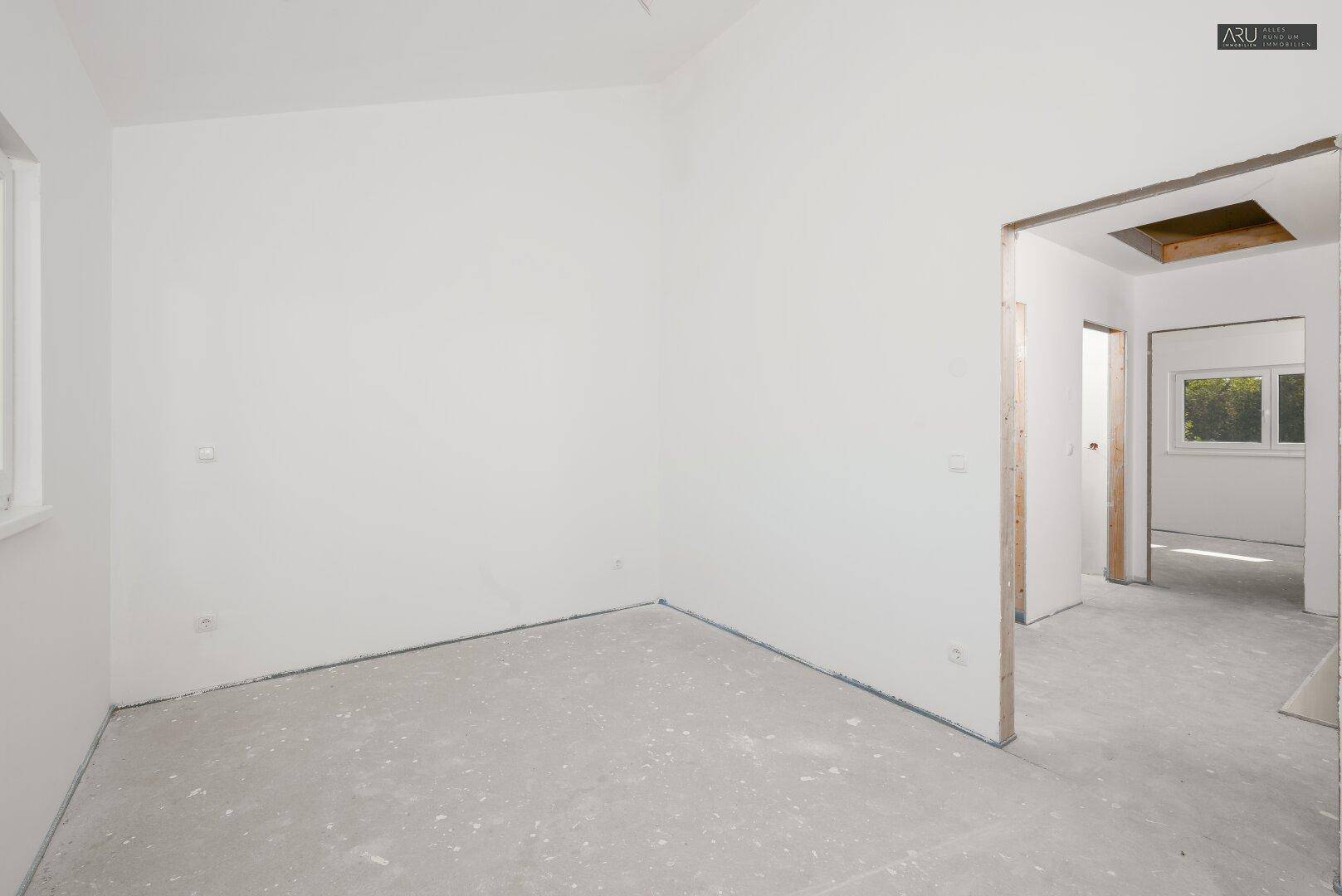 Unfertiges Schlafzimmer mit weißen Wänden, Betonboden und Fenster mit Blick ins Grüne.