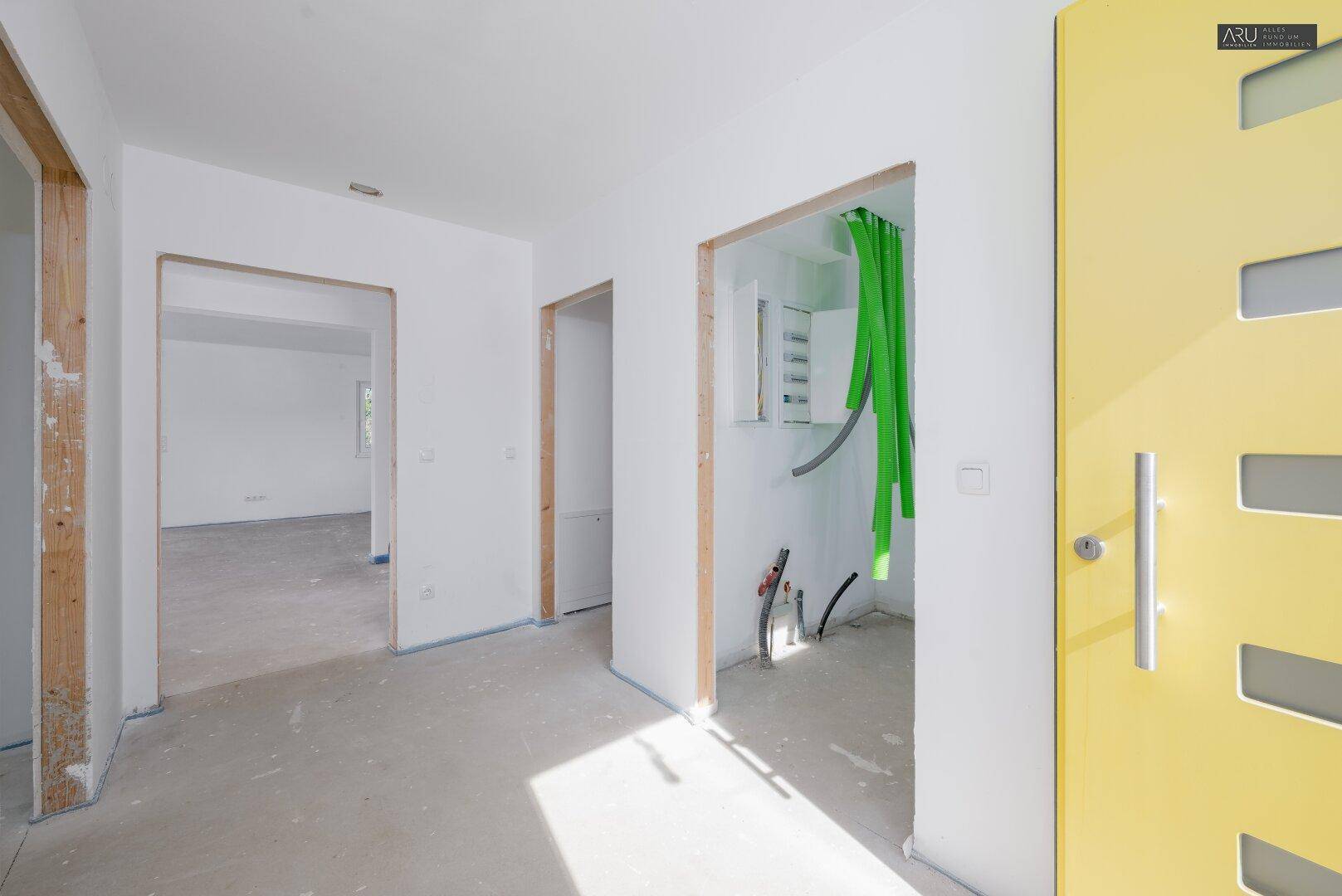Eingangsbereich im Rohbau mit gelber Tür und sichtbaren Installationen für zukünftige Nutzung.
