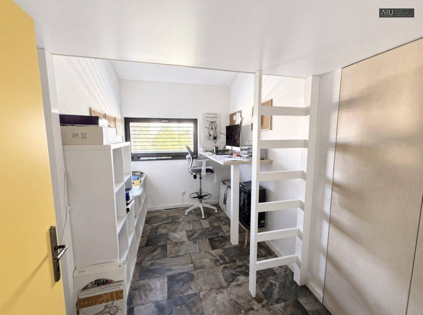 Helles Arbeitszimmer mit Schreibtisch, Regalen und Leiter, gut beleuchtet durch ein Fenster.