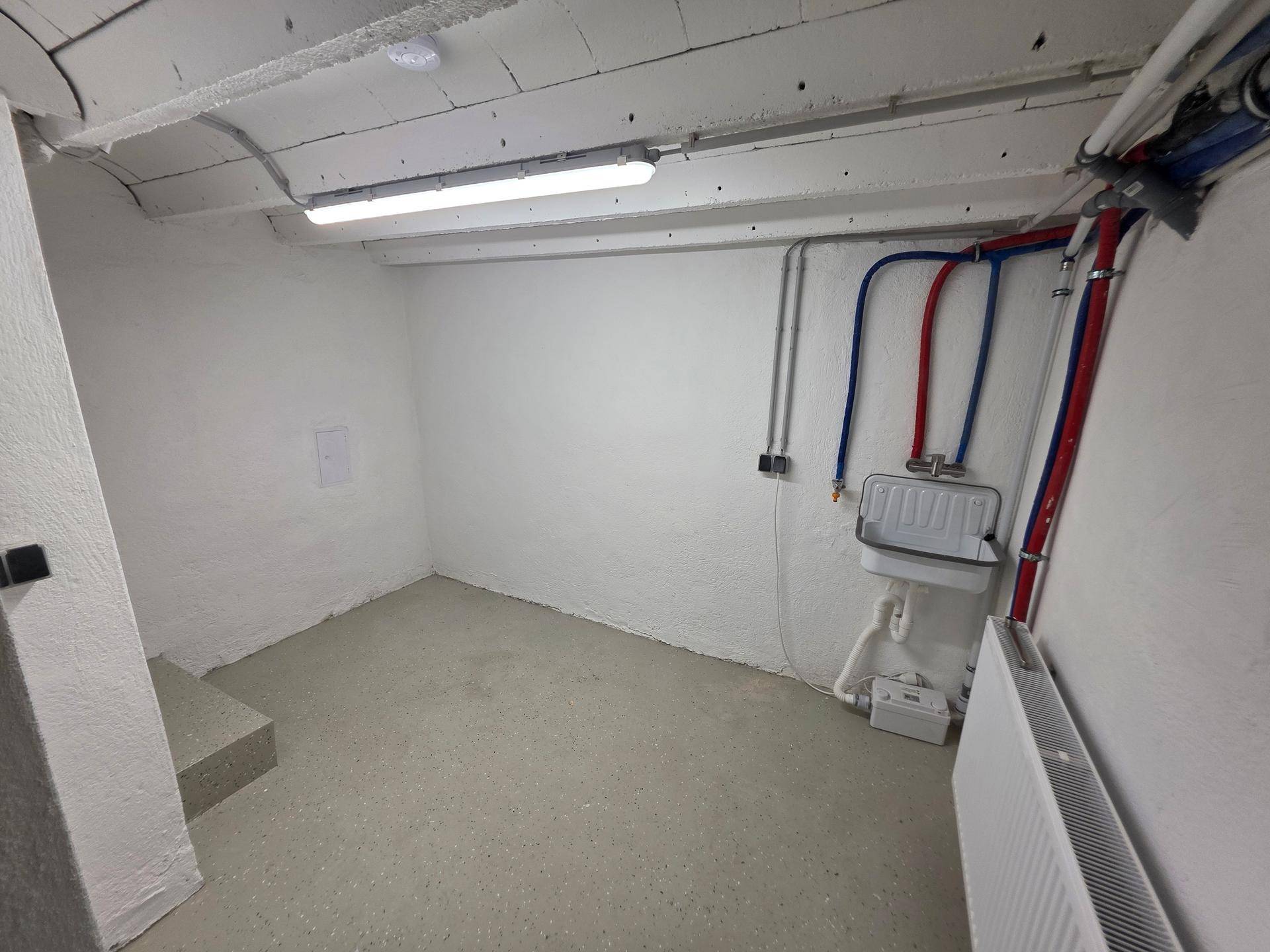 Kellerraum mit Betonboden, weiß gestrichenen Wänden und sichtbaren Versorgungsleitungen.