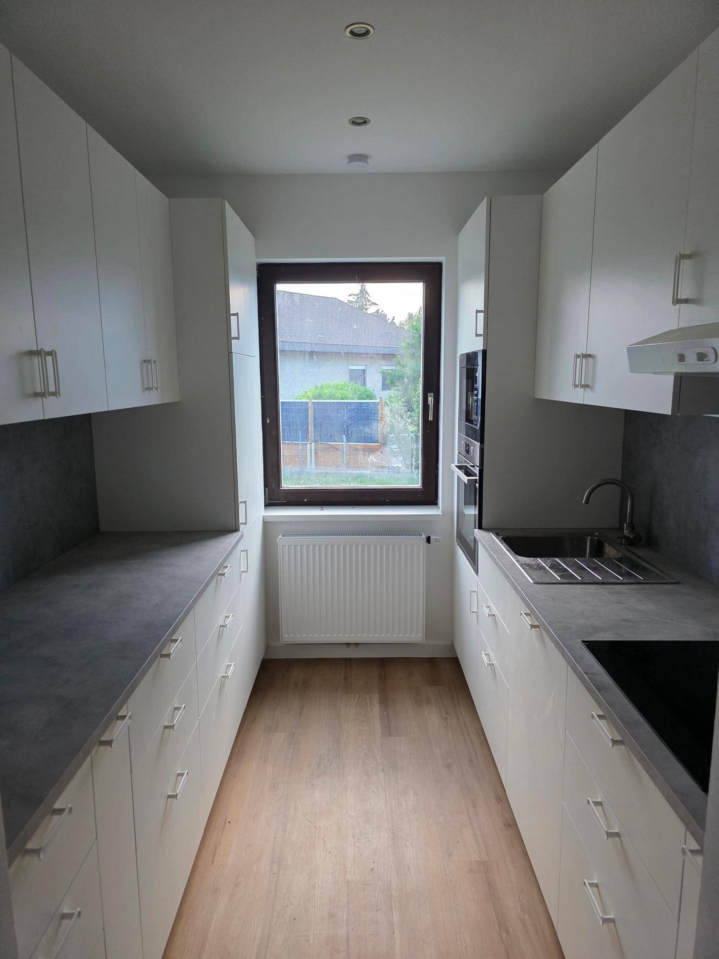 Moderne, schmale Küche mit weißen Schränken, grauen Arbeitsplatten und Fensterblick.