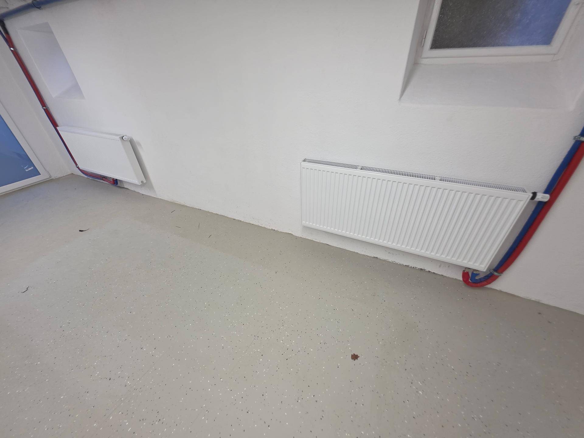 Zwei Heizkörper an einer weißen Wand im Keller, mit sichtbaren Versorgungsleitungen.