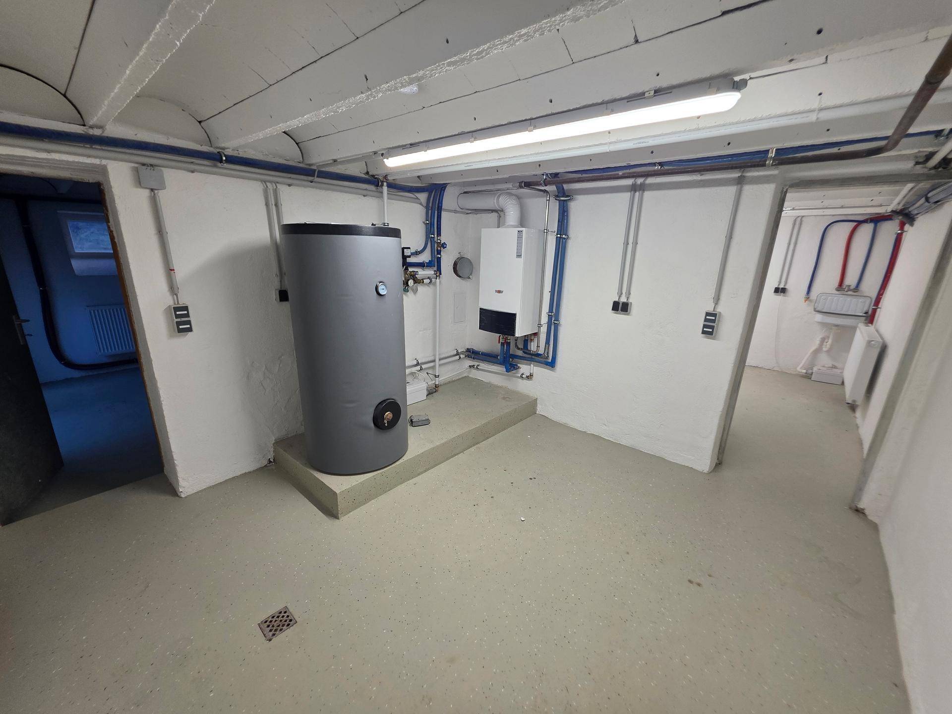Technikraum im Keller mit Heizungsanlage, Warmwasserspeicher und sichtbaren Installationen.