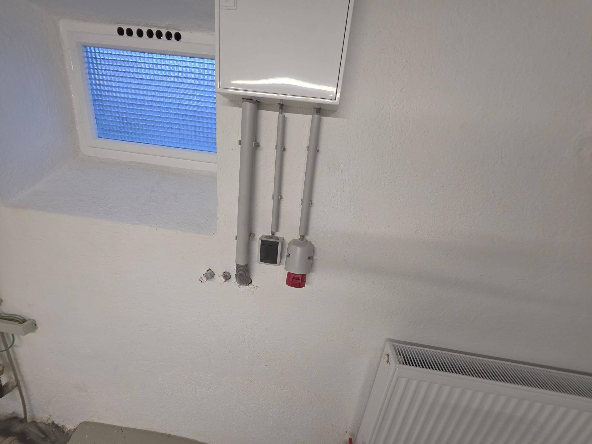 Kellerwand mit Elektroinstallationen, Rohren und einem kleinen Fenster, darunter ein Heizkörper.