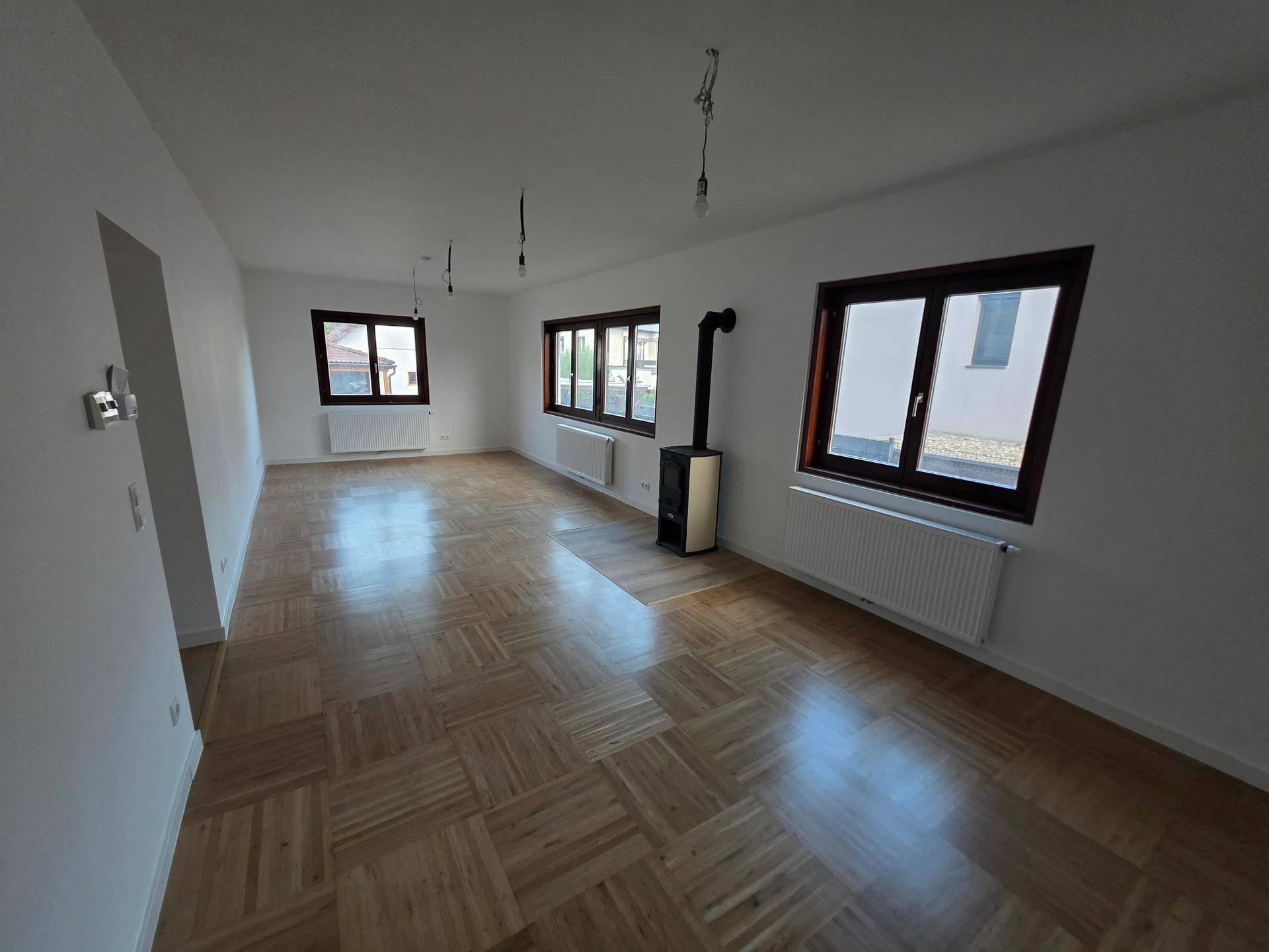 Geräumiges Wohnzimmer mit Parkettboden, mehreren Fenstern und einem Kaminofen.