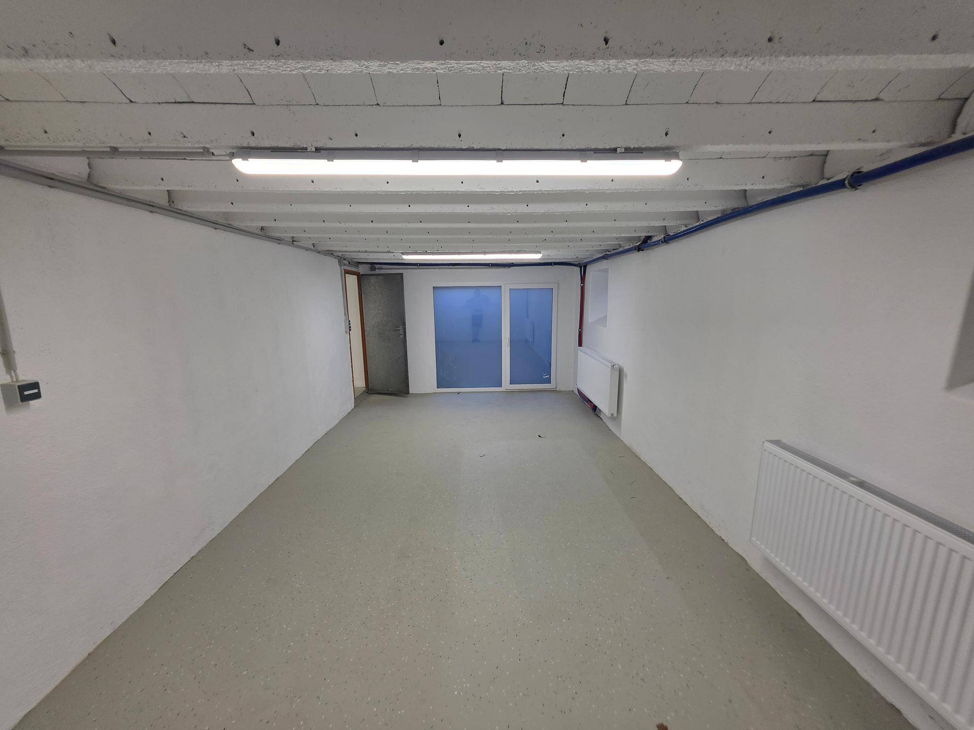 Heller Kellerraum mit Betonboden und großem Fenster, bietet Zugang zum Außenbereich.