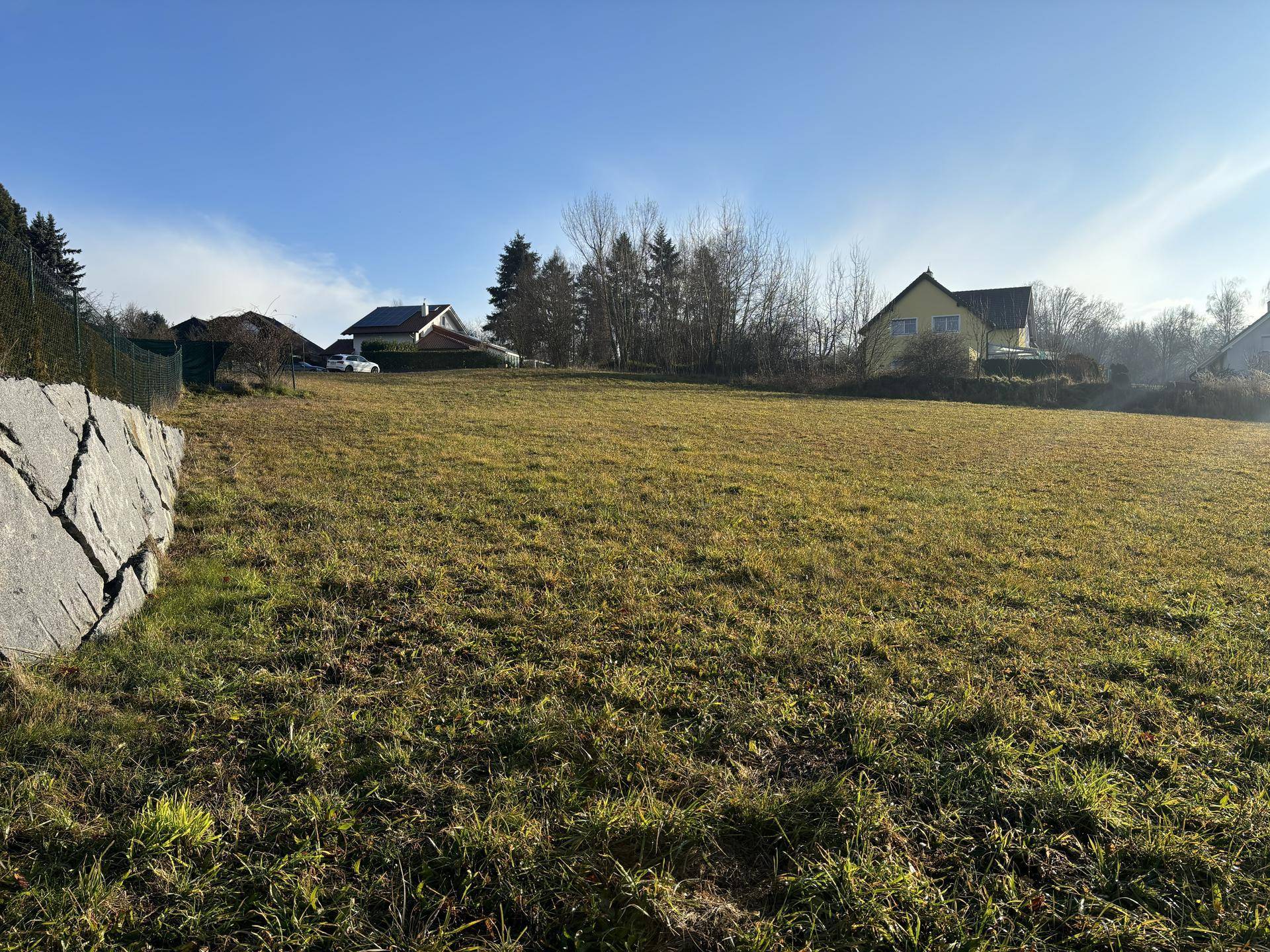 Offenes Feld mit angrenzender Steinmauer und Blick auf weitere Häuser in der Ferne unter klarem Himmel.