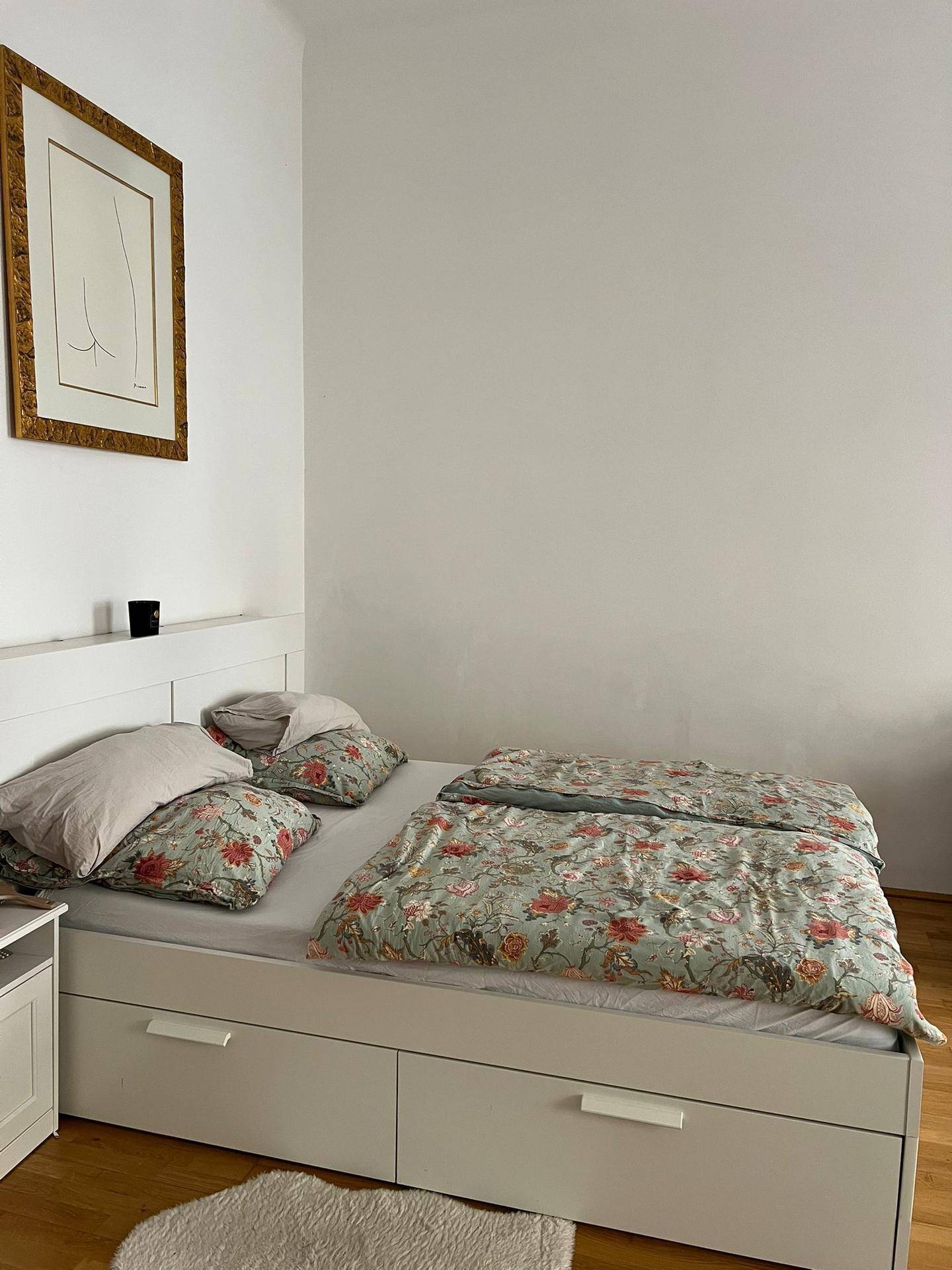 Schlafzimmer mit weißem Bett, floraler Bettwäsche und einem gerahmten Bild an der Wand.