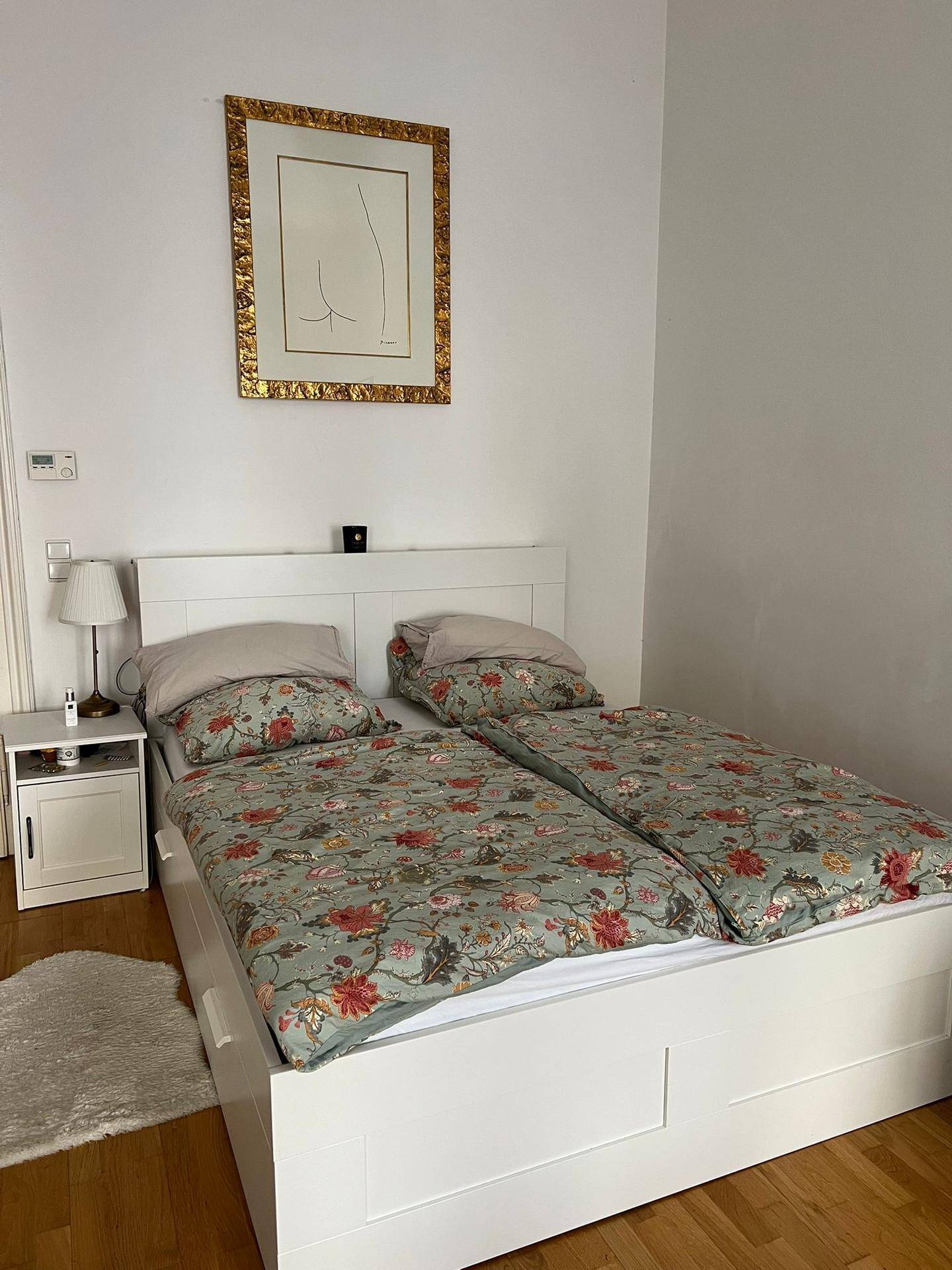 Schlafzimmer mit weißem Bett, floraler Bettwäsche und einem Nachttisch mit Lampe.