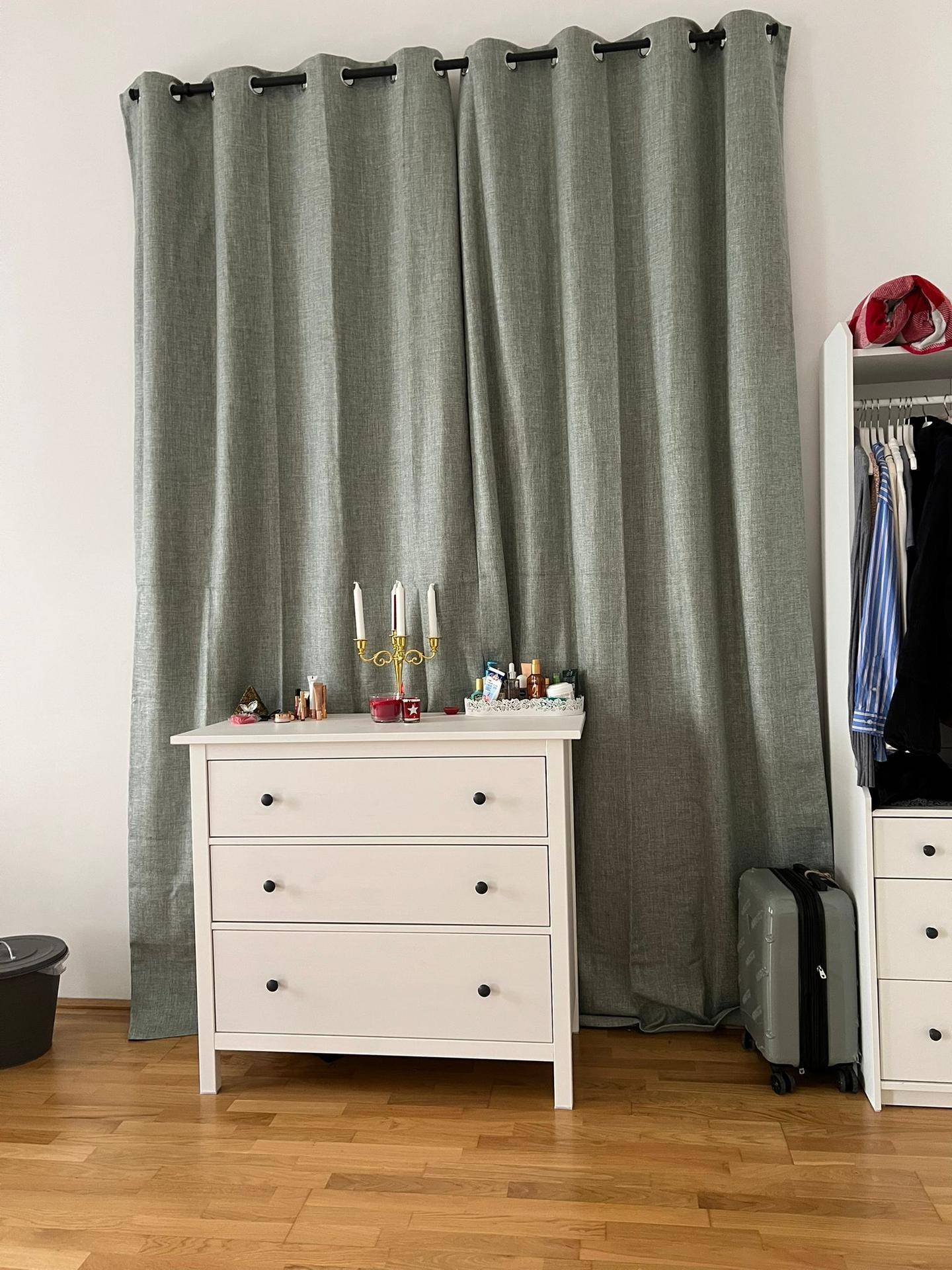 Schlafzimmer mit weißer Kommode, grauen Vorhängen und Holzboden.