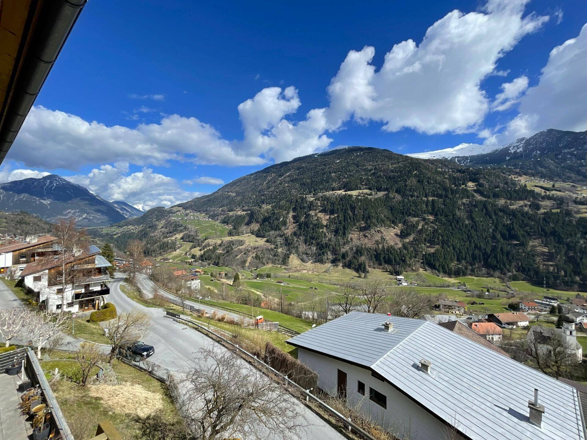 Weitläufiger Blick auf die alpine Landschaft mit bewaldeten Hügeln und einem Tal mit Häusern.