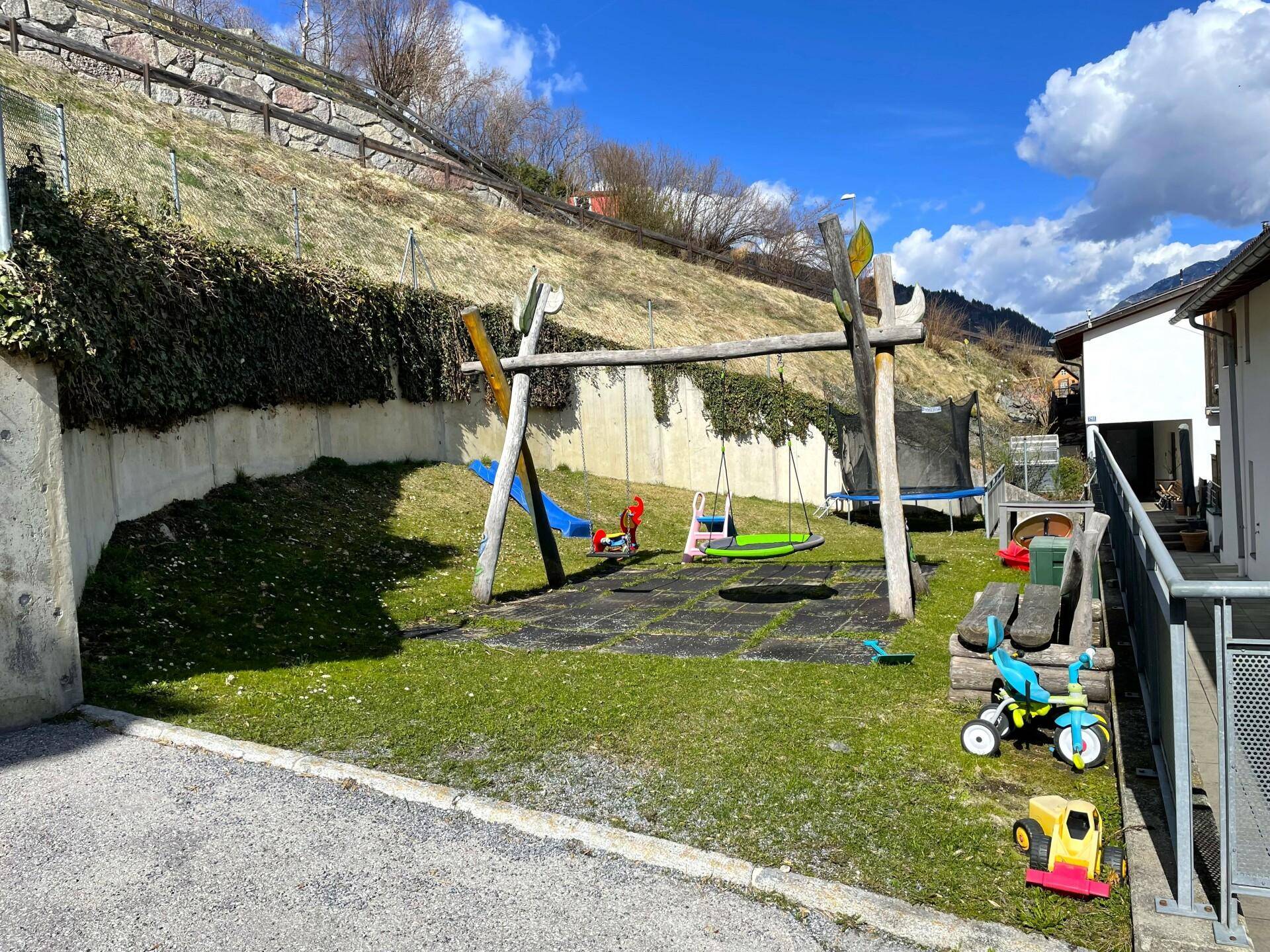 Kleiner Spielplatz mit Schaukeln und Spielgeräten, umgeben von Grünflächen.