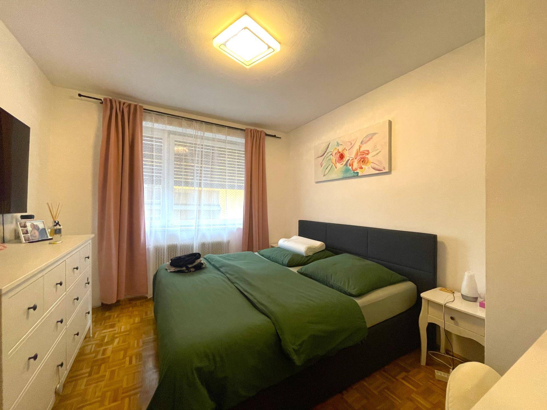 Schlafzimmer mit Doppelbett, Parkettboden und einem Fenster mit Vorhängen.