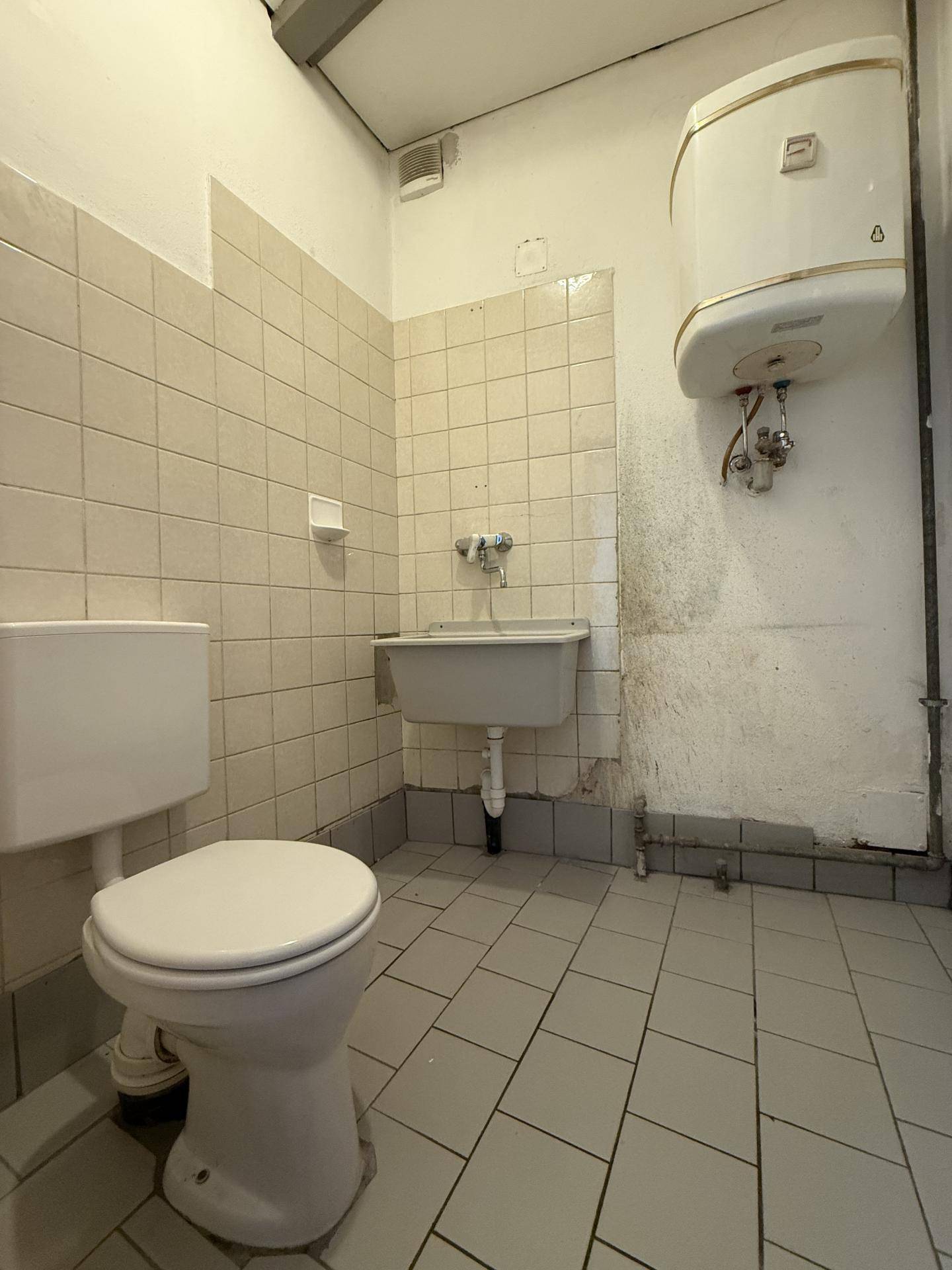 Kleines Badezimmer in Originalzustand mit WC, Waschbecken, Warmwasserboiler und beigefarbenen Fliesen.