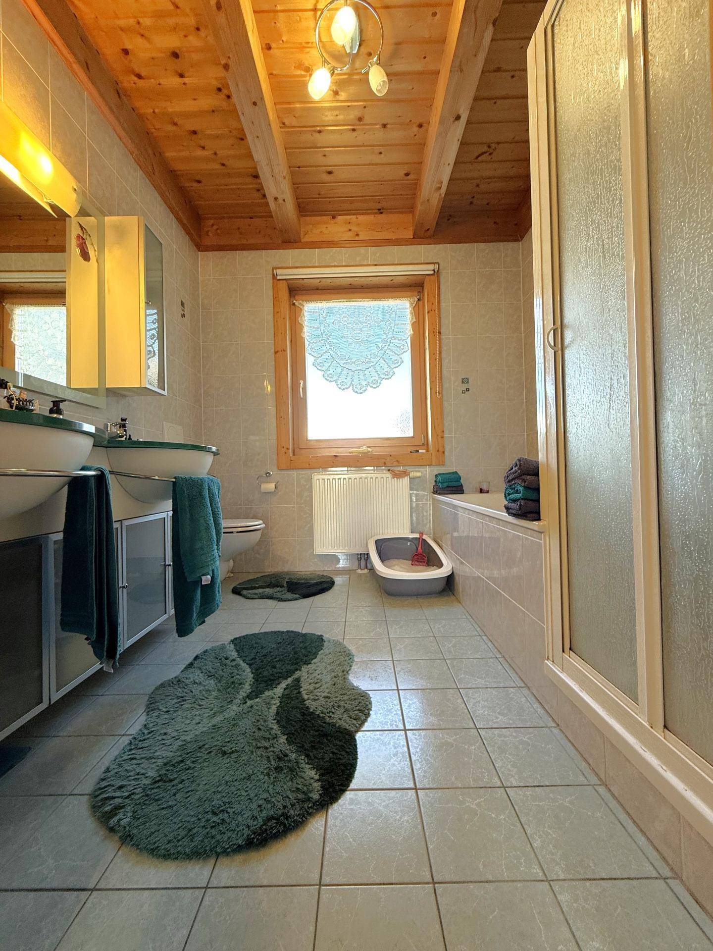 Badezimmer mit Doppelwaschbecken, Dusche und Badewanne, umgeben von hellen Fliesen und Holzbalkendecke.