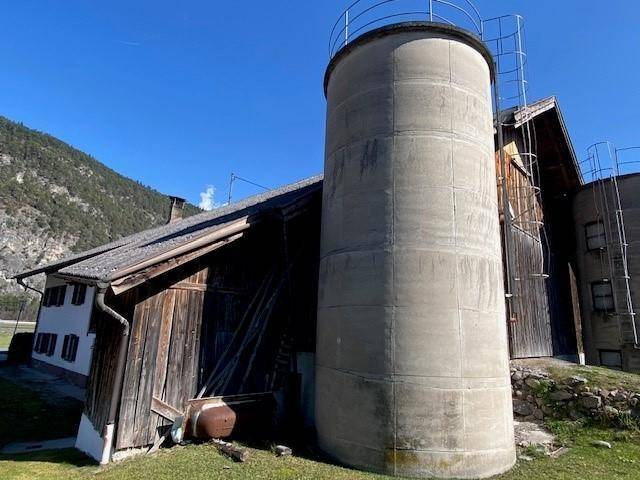 Hohes Betonsilo neben einem rustikalen Holzgebäude unter klarem, blauem Himmel.