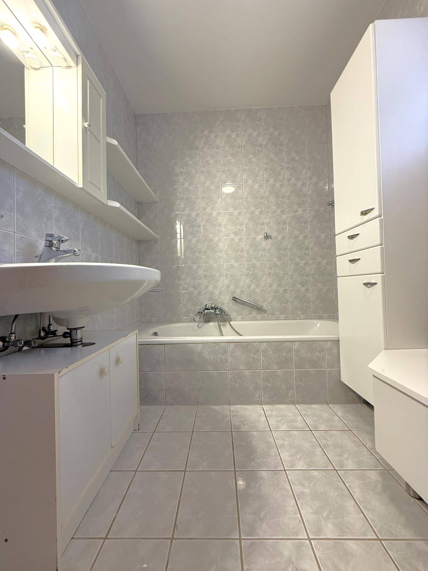 Badezimmer mit hellgrauen Fliesen, einem Waschbecken und einer Badewanne, in gutem Zustand.