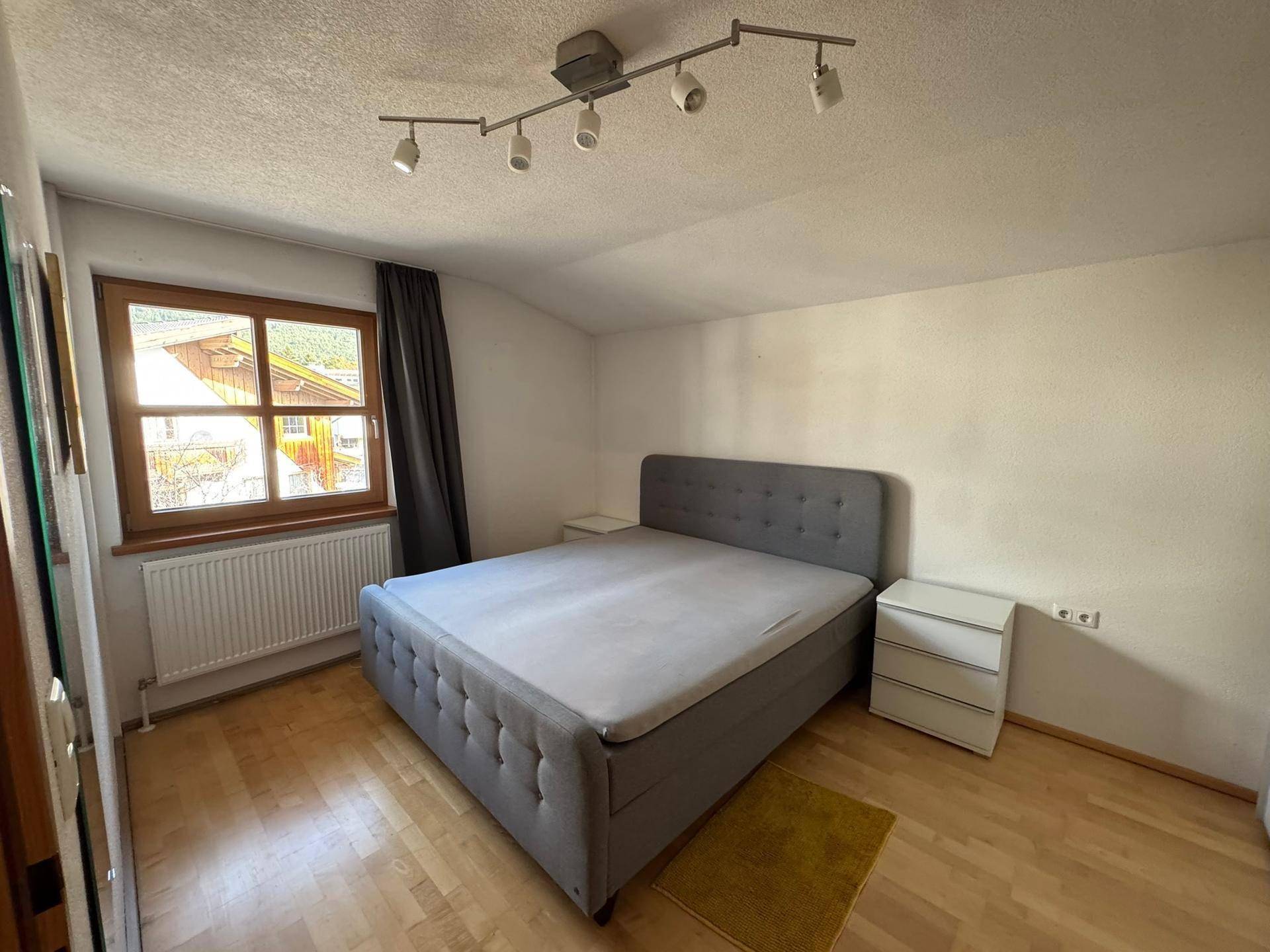 Schlafzimmer mit modernem, grau gepolstertem Bett, Holzboden und einem Fenster.