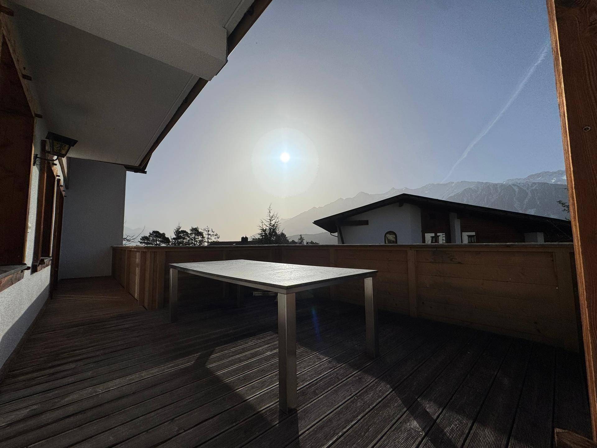 Geräumiger Balkon mit Holzboden, Esstisch und einem weiten Blick auf die Berge bei Sonnenuntergang.