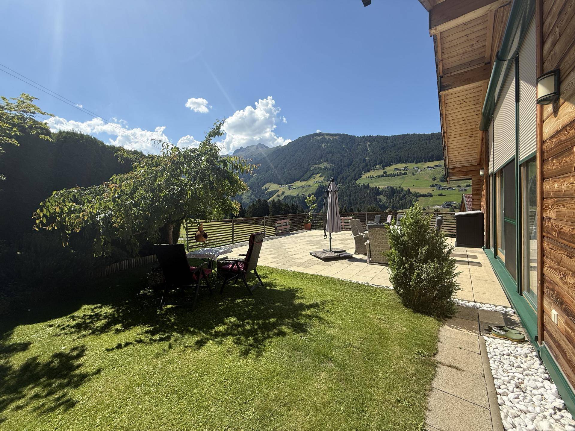 Sonniger Gartenbereich mit Rasenfläche, Bäumen und einer Terrasse mit Bergblick.