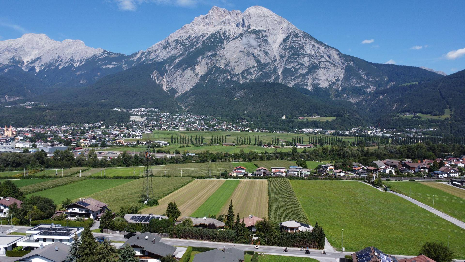 Weitläufige Luftaufnahme einer Siedlung mit Feldern und einem imposanten Bergmassiv im Hintergrund.