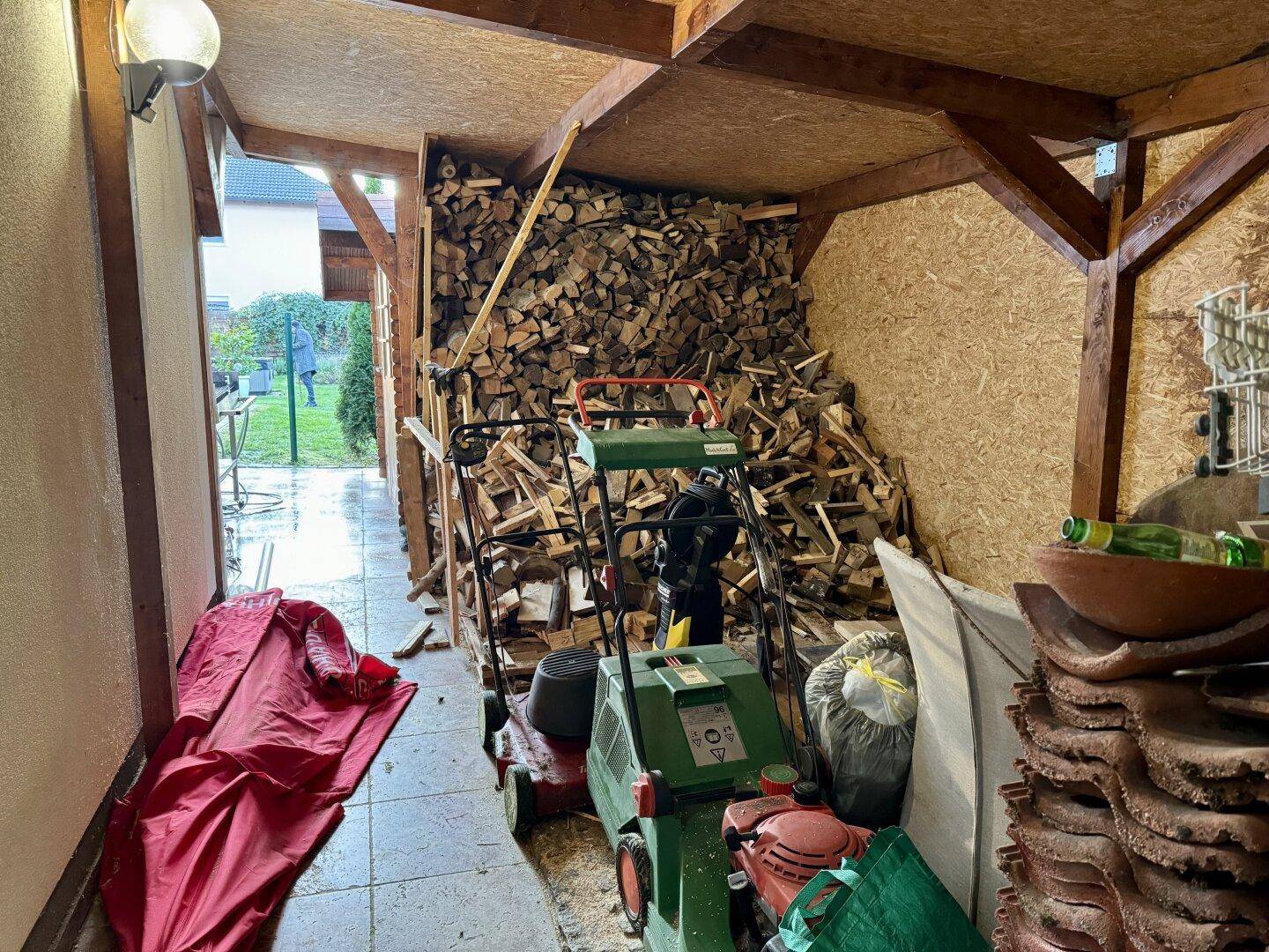 Überdachter Holzschuppen mit Brennholzstapel und Gartengeräten.