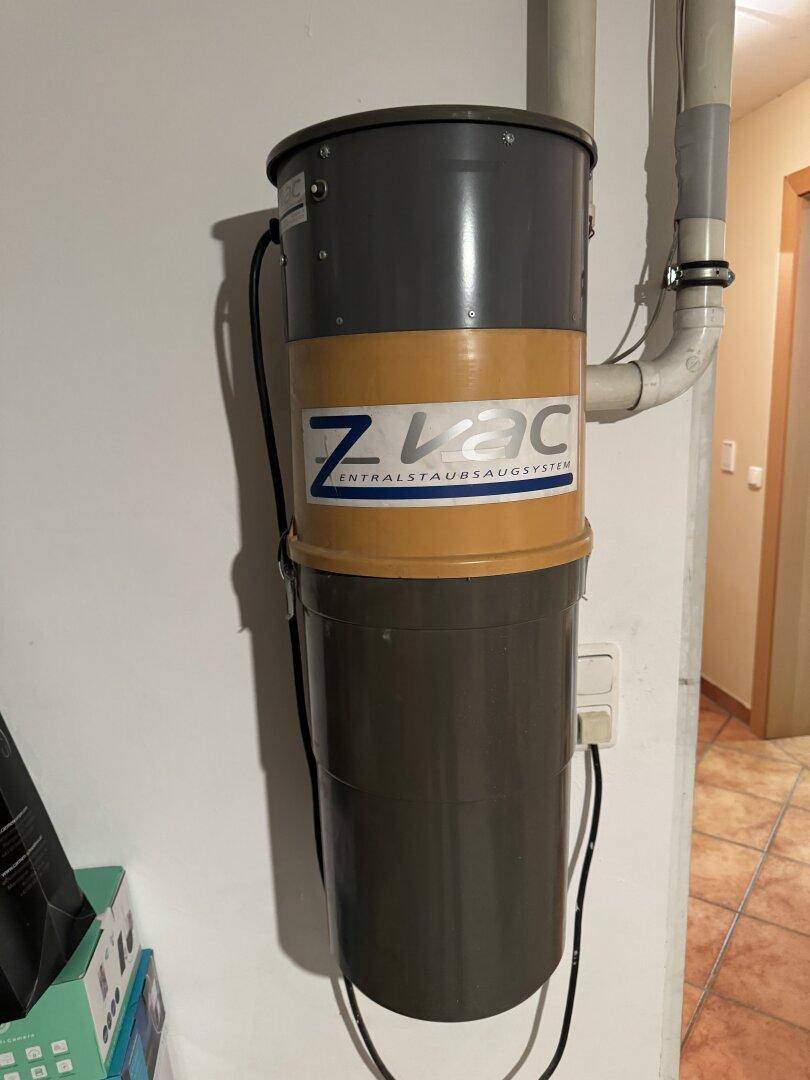 An der Wand montiertes Zentralstaubsaugsystem mit angeschlossenen Rohren und Fliesenboden in einem Hauswirtschaftsraum.
