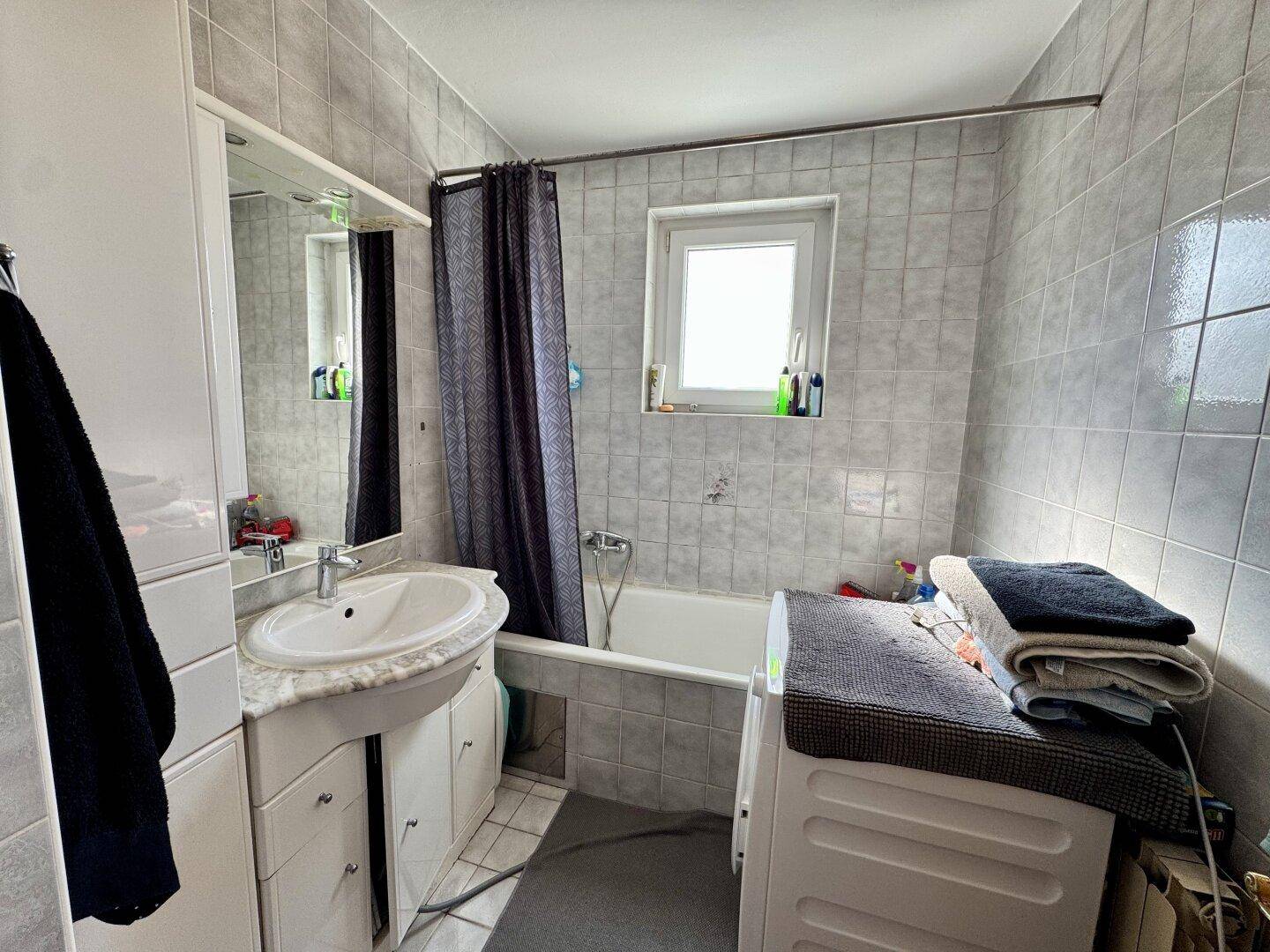Badezimmer mit Badewanne, Waschbecken und Fenster, ausgestattet mit grauen Fliesen.