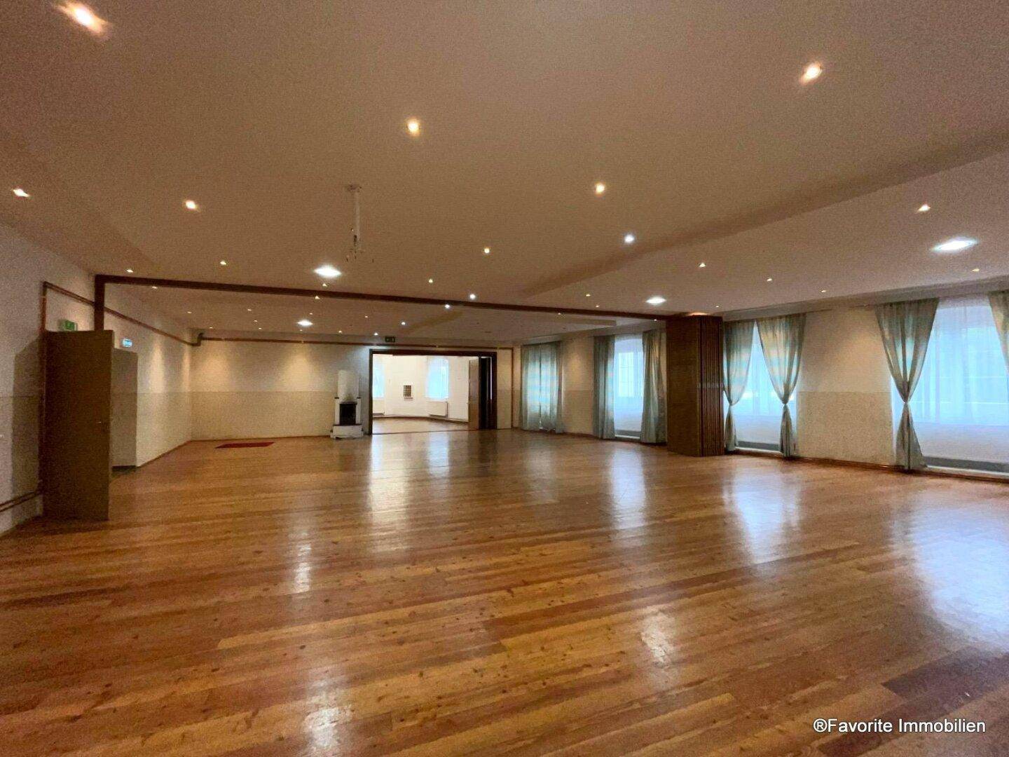 Weitläufiger, leerer Veranstaltungssaal mit Holzboden, vielen Fenstern und moderner Deckenbeleuchtung, sehr geräumig.