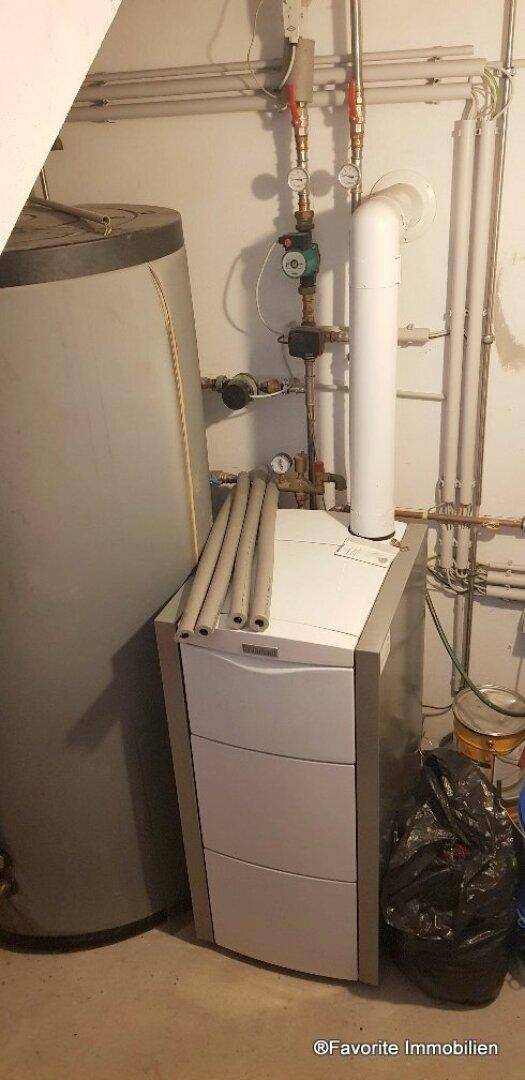 Heizungsraum mit Warmwasserspeicher und Heizkessel, umgeben von Rohren und Installationen.
