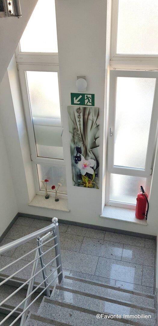 Treppenaufgang mit großen Fenstern, einem Feuerlöscher und einem dekorativen Bild.
