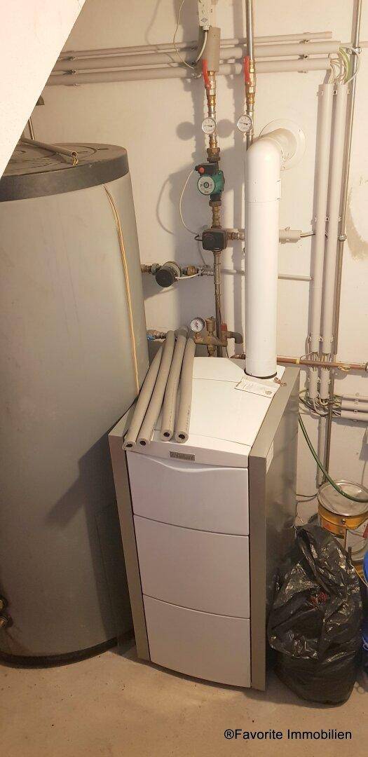 Heizungsraum mit Warmwasserspeicher und Heizkessel, umgeben von Rohren und Installationen.