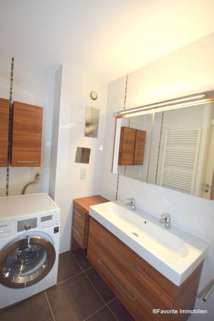 Modernes Badezimmer mit Waschbeckenunterschrank, Waschmaschine und Fliesenboden, inklusive Spiegel und Beleuchtung.